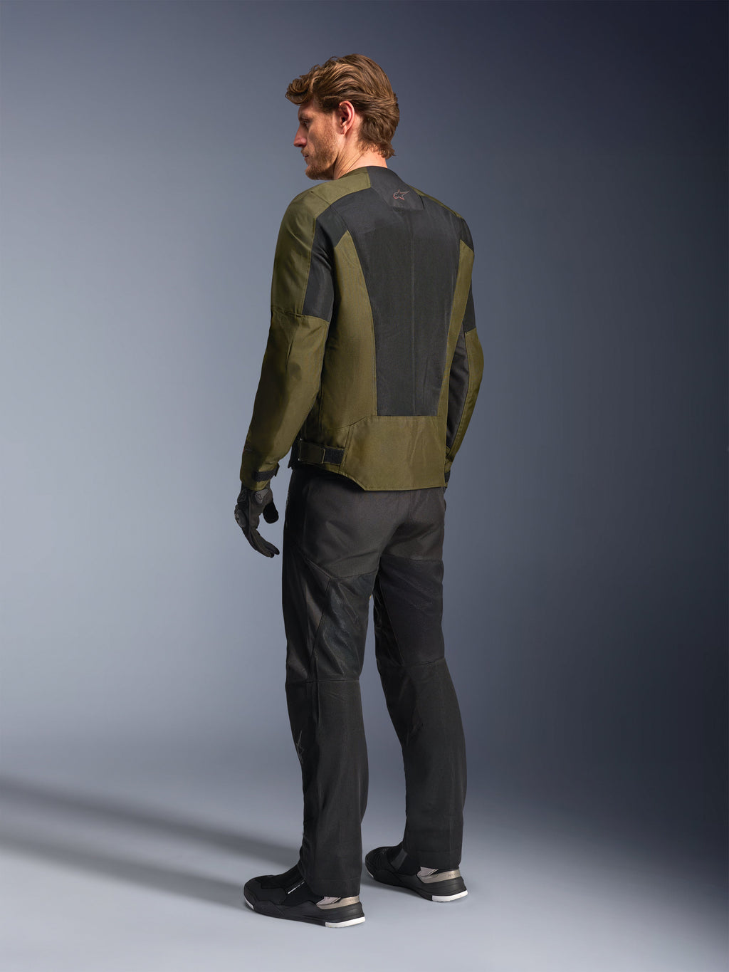 Mannelijk model draagt Alpinestars C-1 Air Jacket in Khaki, urban motorjas, achteraanzicht met nadruk op de grote zwarte mesh-panelen op de rug, gecombineerd met een zwarte technische motorbroek en handschoenen, eigentijdse motoruitrusting voor warm weer
