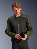 Man die de Alpinestars C-1 Air Jacket in Khaki draagt, urbane motorjas met olijfgroen textiel en zwarte mesh-panelen, kijkt naar beneden terwijl hij zwarte motorhandschoenen vasthoudt, strakke en eigentijdse stijl voor rijden op de weg.