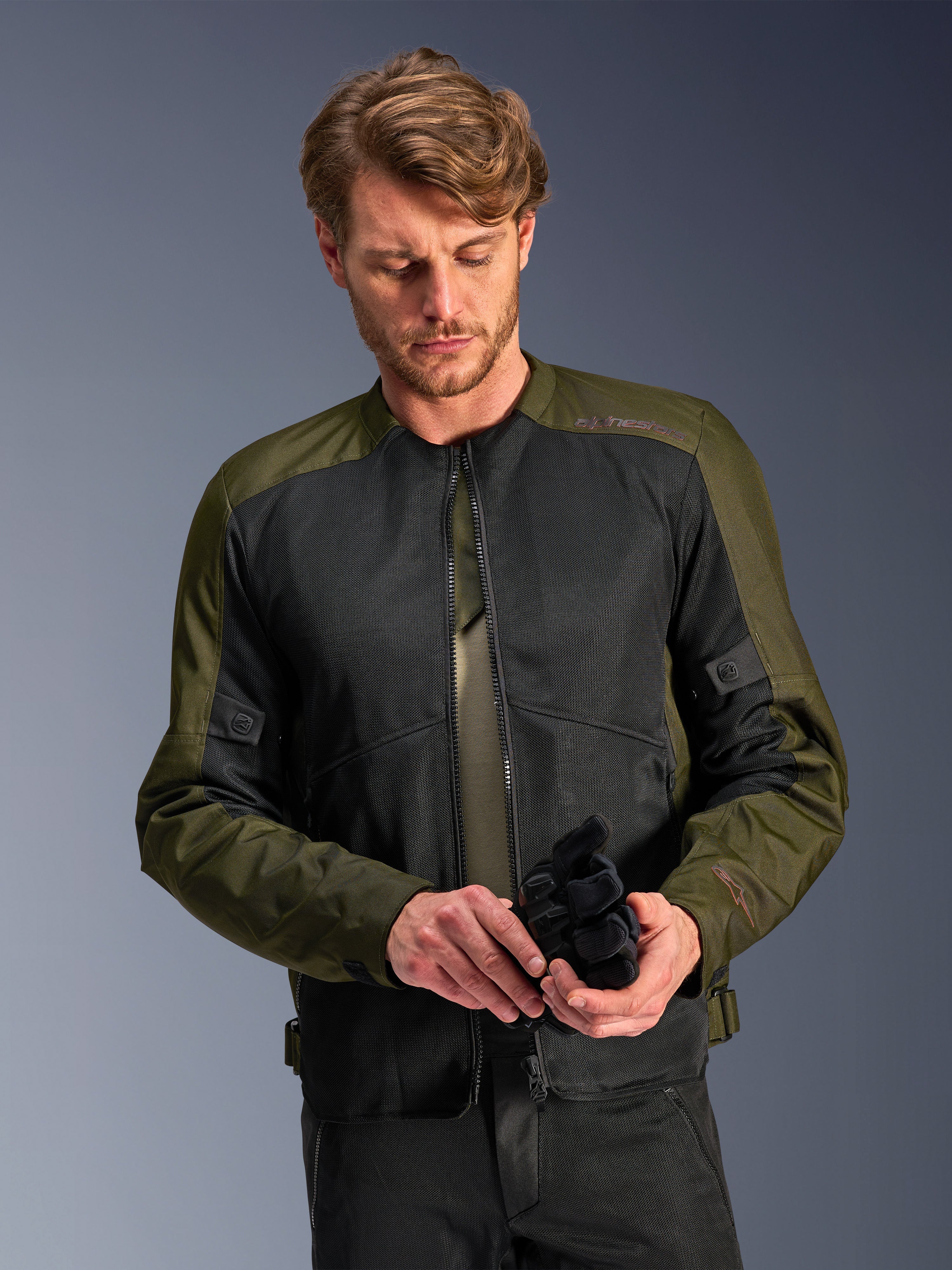 Man die de Alpinestars C-1 Air Jacket in Khaki draagt, urbane motorjas met olijfgroen textiel en zwarte mesh-panelen, kijkt naar beneden terwijl hij zwarte motorhandschoenen vasthoudt, strakke en eigentijdse stijl voor rijden op de weg.