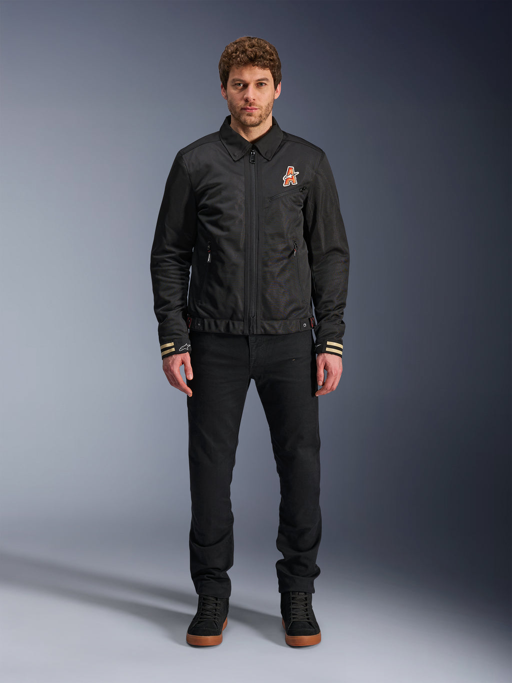 Mannelijk model draagt de zwarte Alpinestars Ignite Air Jacket, voorzien van een klassieke overhemdkraag, een volledige ritssluiting aan de voorzijde en een oranje logo op de borst. Gecombineerd met een zwarte broek en zwarte hoge sneakers voor een eigentijdse urban motorlook.