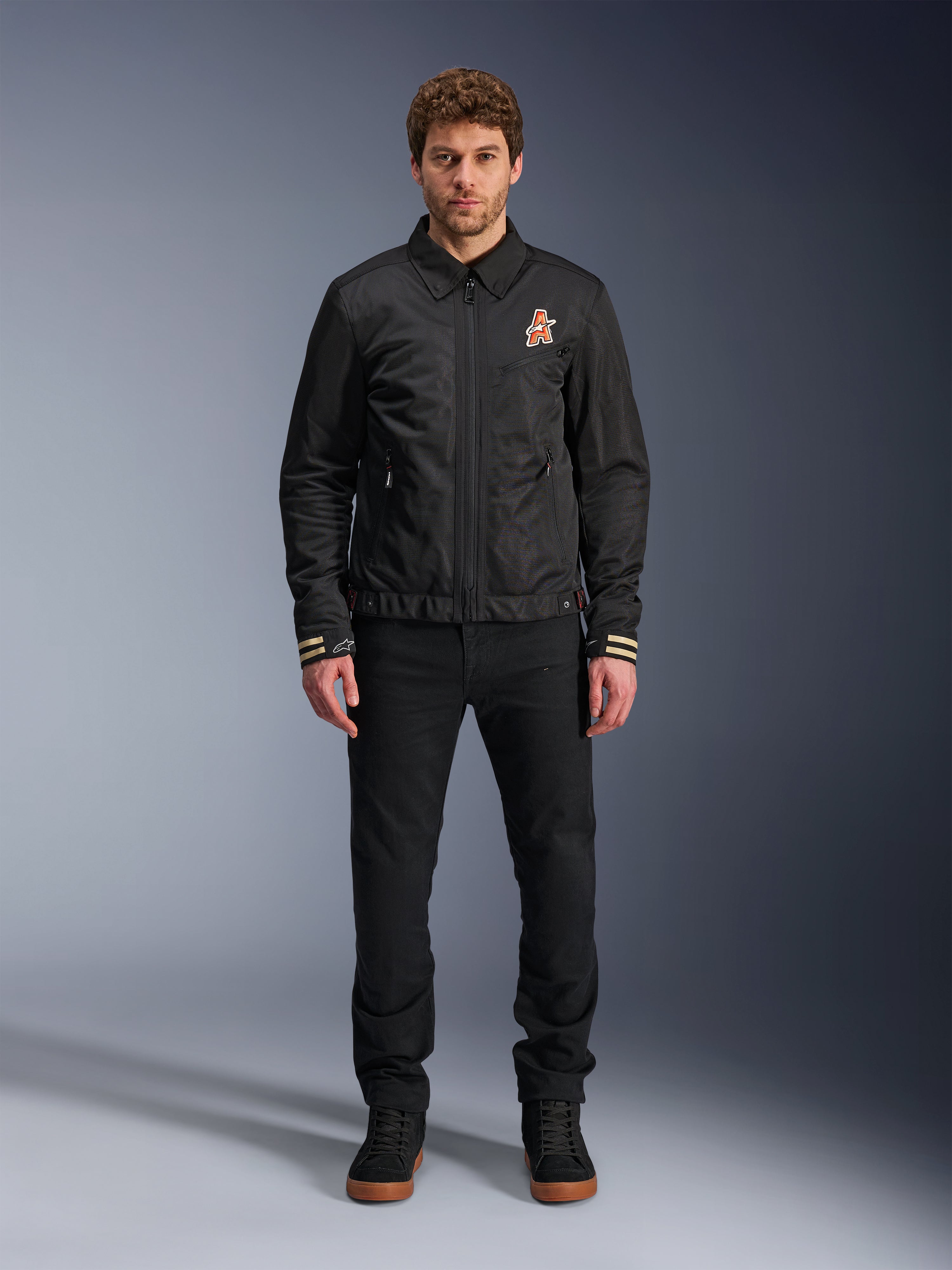 Mannelijk model draagt de zwarte Alpinestars Ignite Air Jacket, voorzien van een klassieke overhemdkraag, een volledige ritssluiting aan de voorzijde en een oranje logo op de borst. Gecombineerd met een zwarte broek en zwarte hoge sneakers voor een eigentijdse urban motorlook.