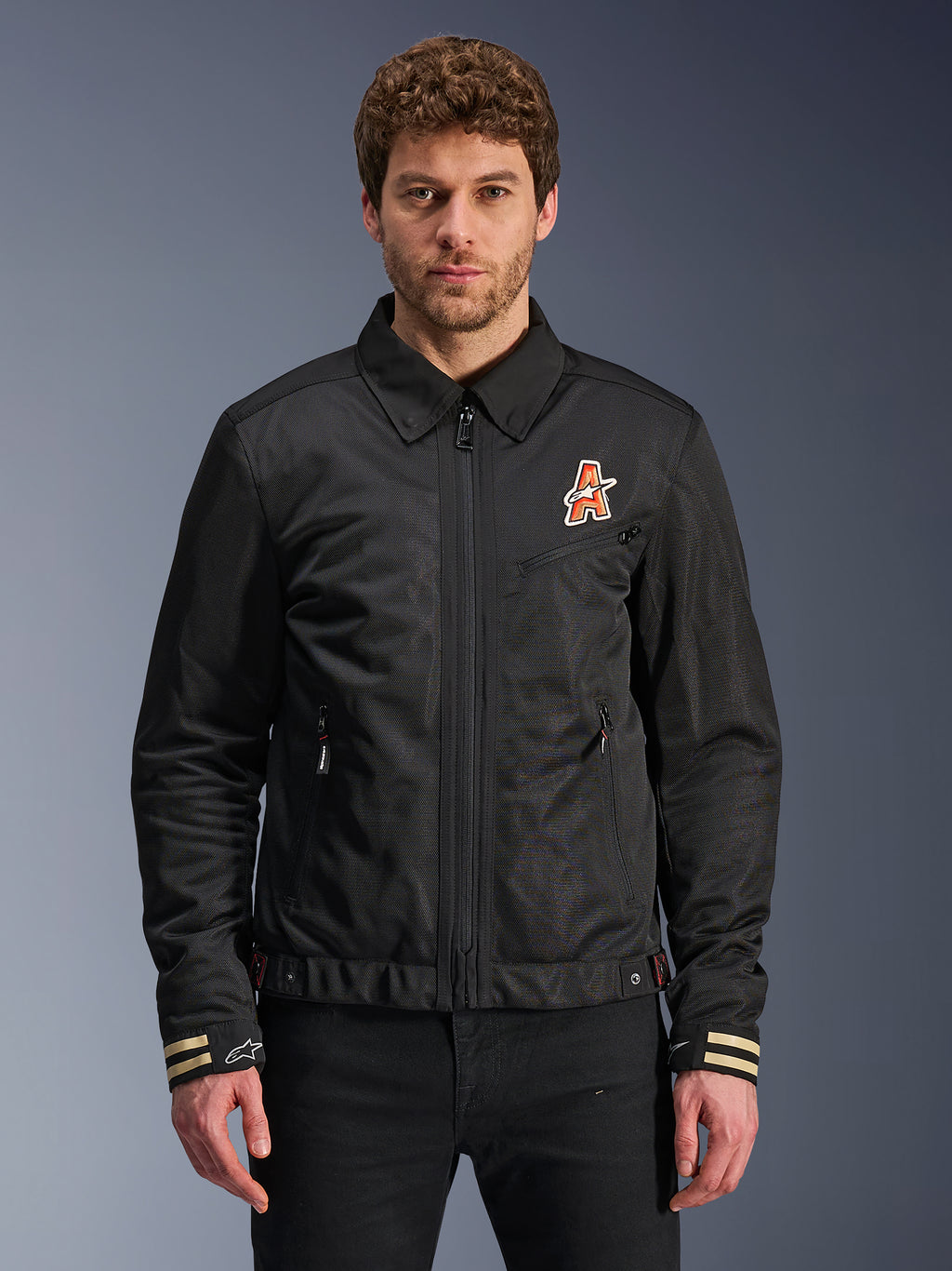 Alpinestars Ignite Air Jacket, zwarte motorjas, traditionele overhemdkraag, oranje en wit 'A'-logo op de borst, beige gestreepte manchetten met wit sterlogo, urban contemporary motorkleding