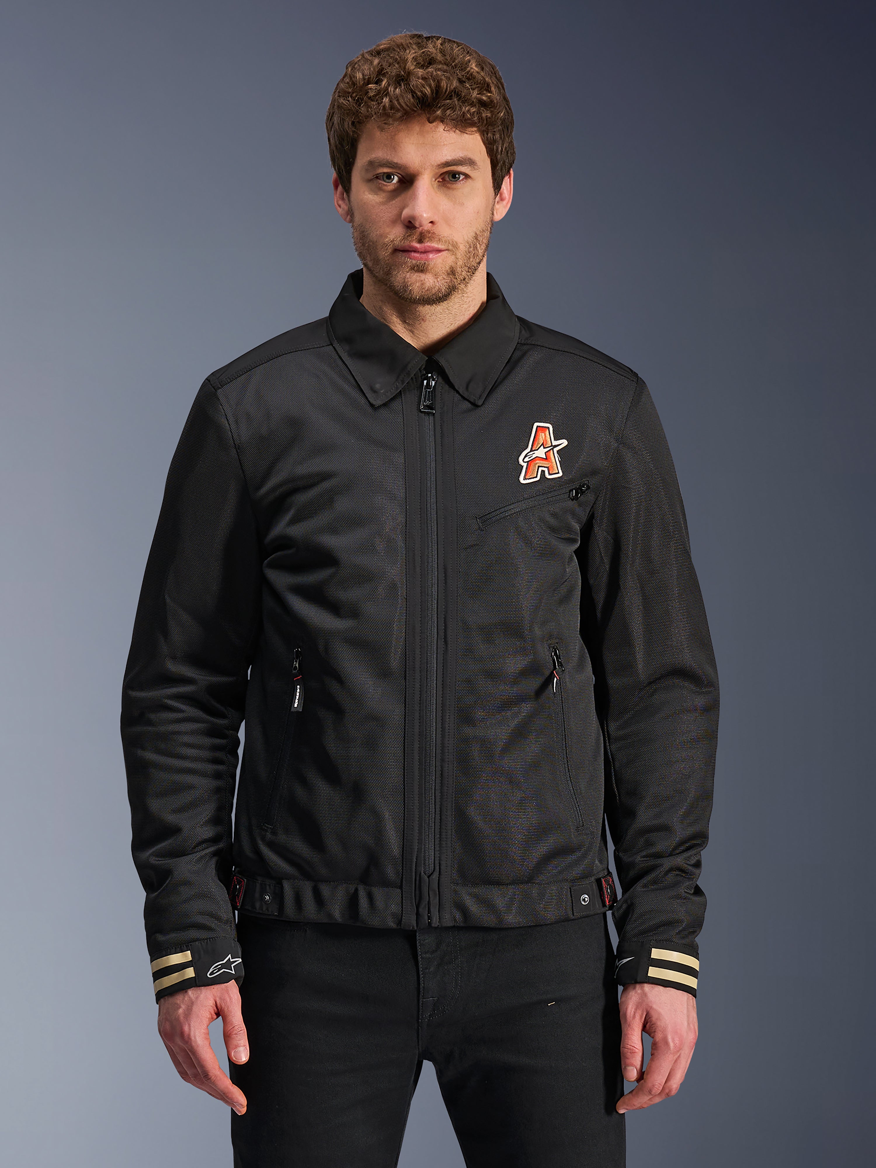 Alpinestars Ignite Air Jacket, zwarte motorjas, traditionele overhemdkraag, oranje en wit 'A'-logo op de borst, beige gestreepte manchetten met wit sterlogo, urban contemporary motorkleding