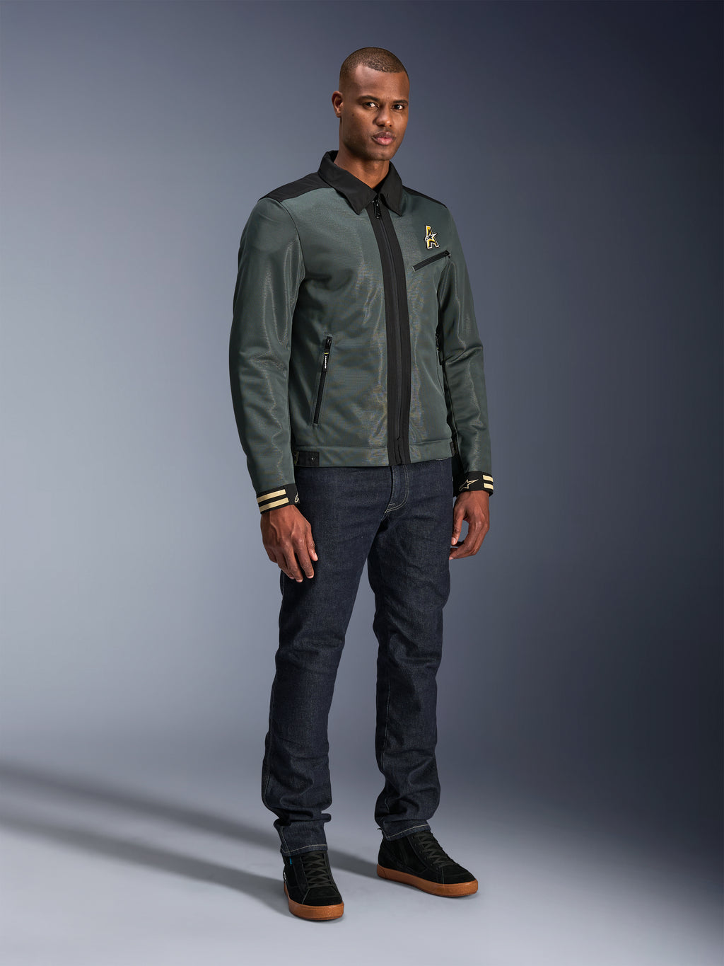 Alpinestars Ignite Air Jacket, Urban Gray Black Blue, urban motorjas, gedragen door een model met donkere denim jeans en zwarte sneakers, voorzien van een mesh body, traditionele overhemdkraag, Harrington-geïnspireerd silhouet en gouden Alpinestars branding.