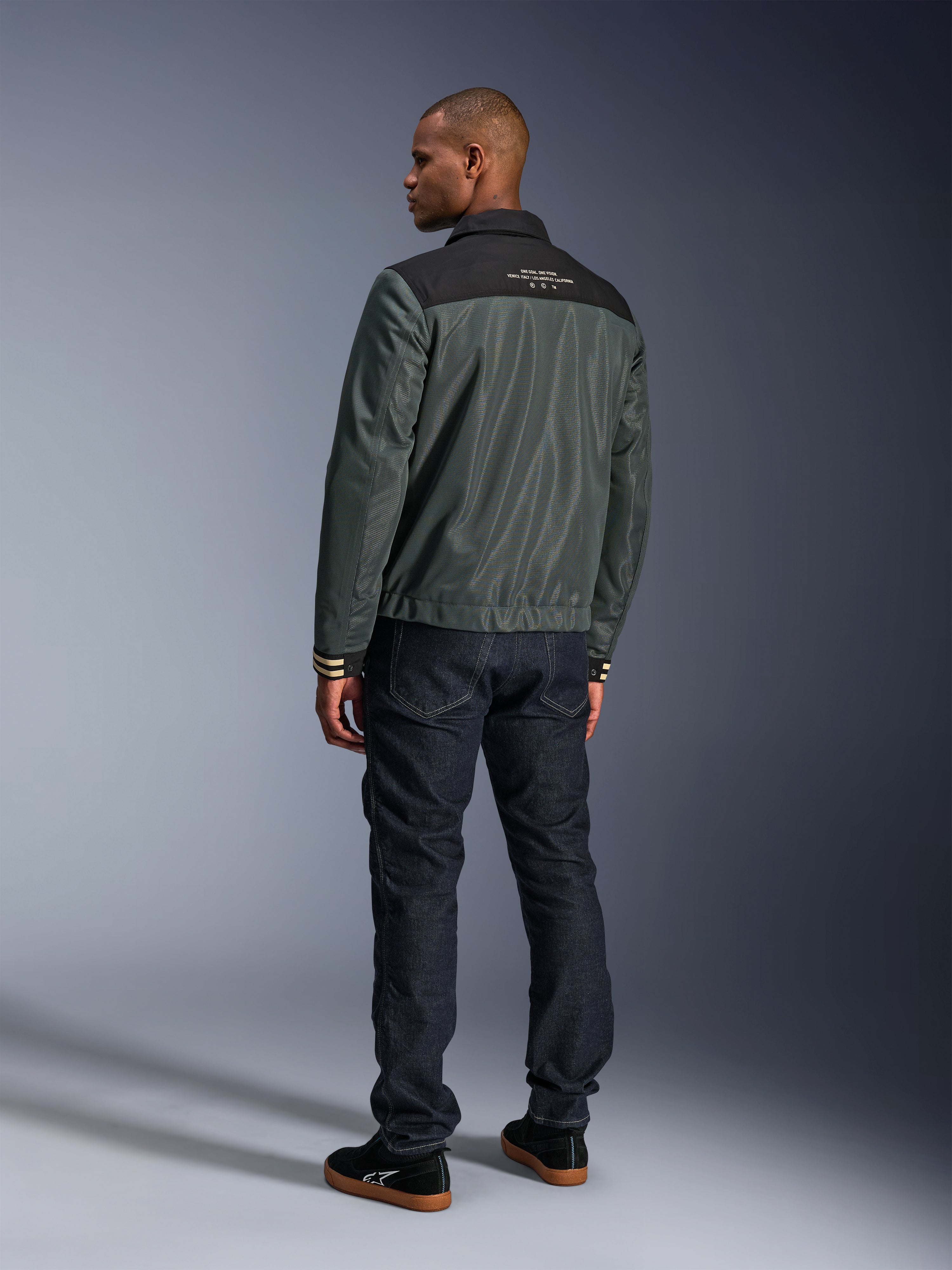 Alpinestars Ignite Air Jas in Urban Gray Black Blue, achteraanzicht op model, met een mesh body, een zwart schouderstuk met witte tekst en gestreepte manchetten, gecombineerd met een donkere denim jeans en zwarte sneakers, urban lifestyle motorkleding