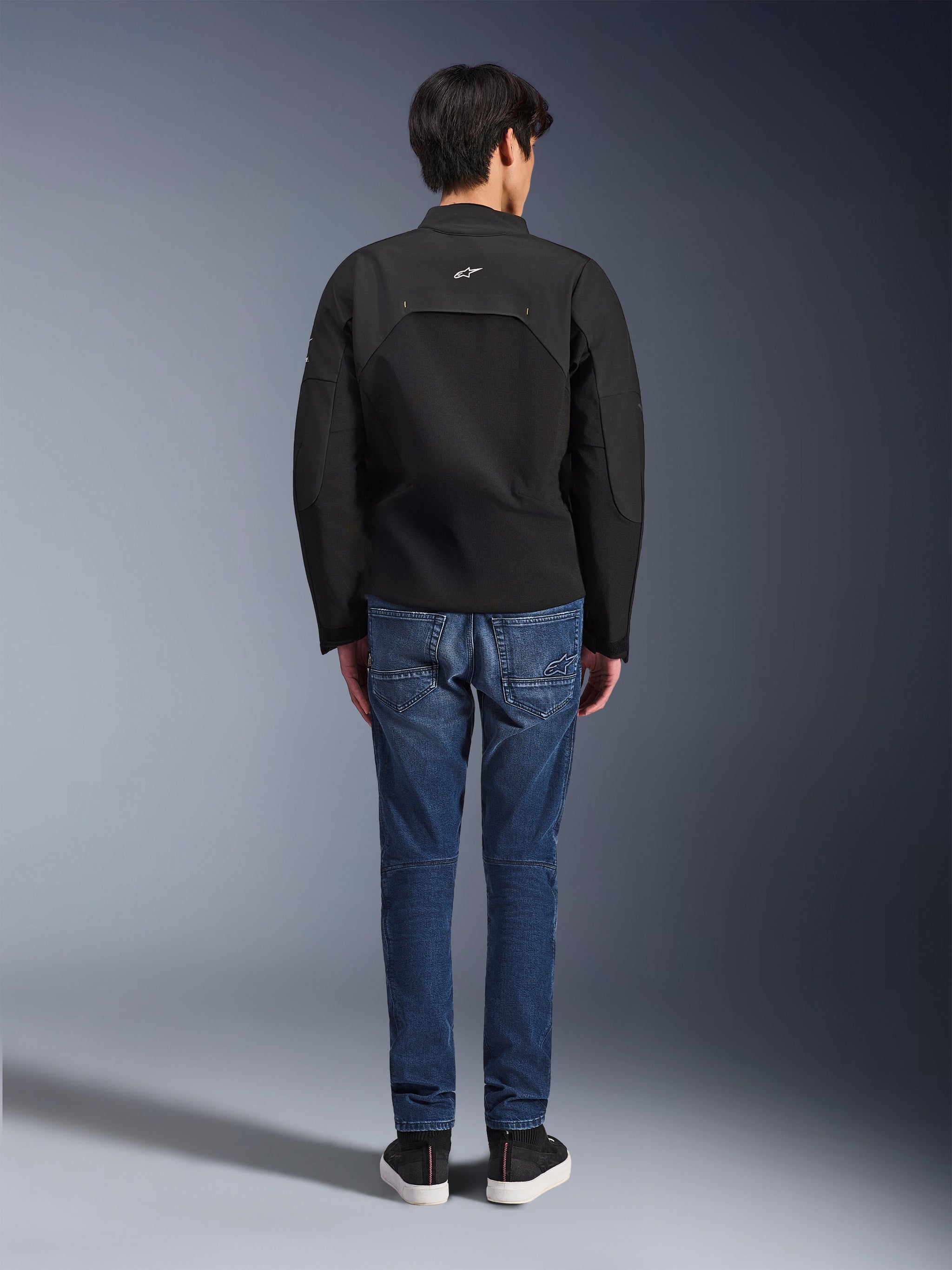 Achteraanzicht van een model met de Alpinestars Provoke Jacket, zwarte technische textiel motorjas, gestyled met een blauwe denim motorjeans en zwarte sneakers, toont de achterzijde en een klein wit Alpinestars-logo, urban eigentijds ontwerp