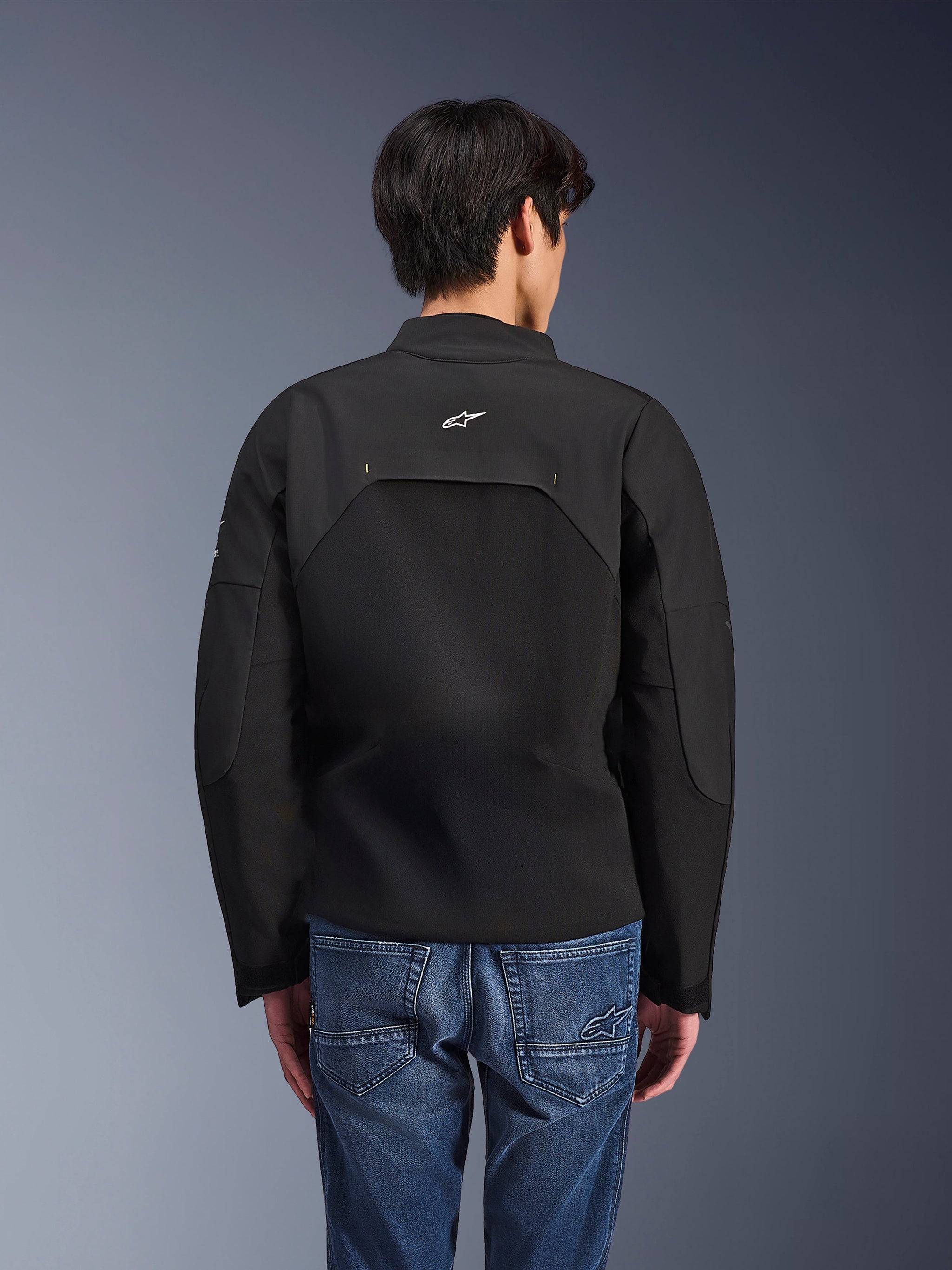 Alpinestars Provoke Jas, stedelijke motorjas, zwart, achteraanzicht met wit Astars-logo en technische ripstop-panelen, gedragen met blauwe denim jeans, eigentijds ontwerp voor woon-werkverkeer