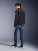 Alpinestars Provoke Jacket, zwarte urban motorjas, gedragen door een model in driekwart achteraanzicht, gecombineerd met blauwe denim jeans en zwarte sneakers, voorzien van ripstop softshell-inzetstukken en witte Alpinestars-branding op de mouw en rug.
