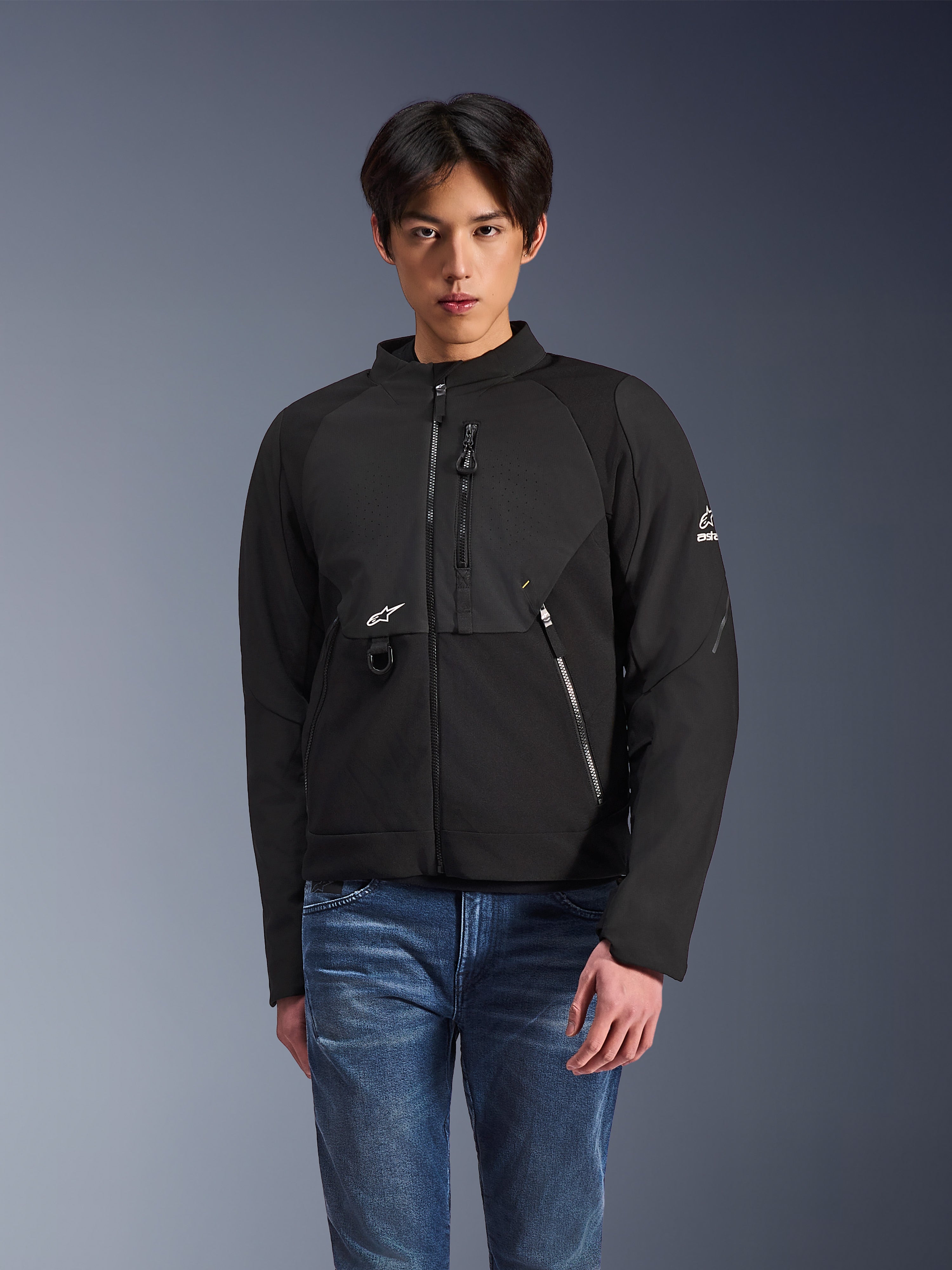 Alpinestars Provoke Jacket, zwarte textiele urban motorjas, gedragen door een mannelijk model met blauwe jeans, voorzien van laser-geperforeerde borstpanelen, meerdere zakken met ritssluiting, wit A-star logo op de borst en mouw, strak modern ontwerp voor ritten op de weg