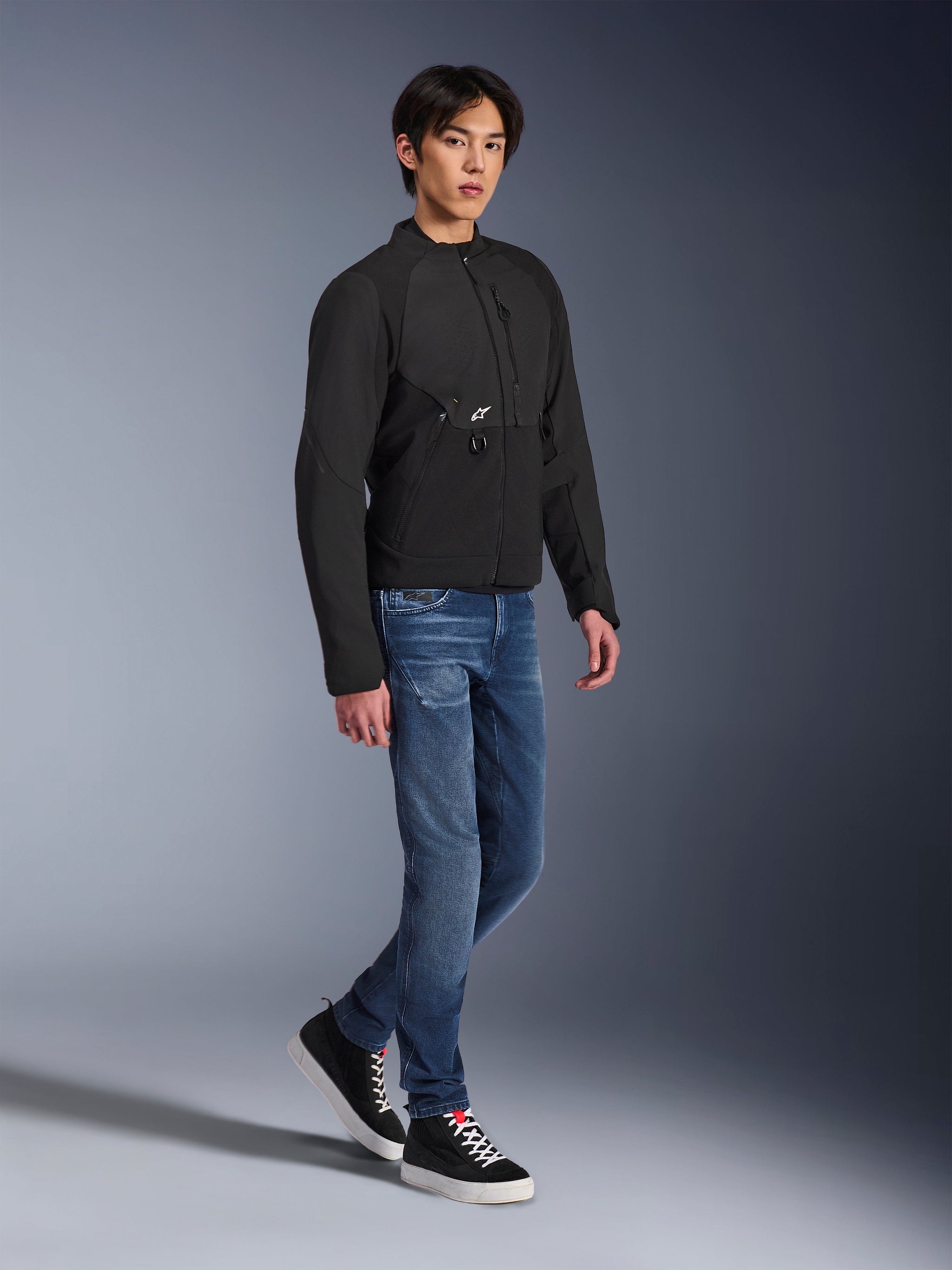 Alpinestars Provoke Jas, stedelijke motorjas, zwart, gedragen door een mannelijk model met blauwe jeans en zwarte sneakers, technisch textielontwerp met een klein Alpinestars-logo, full-body lifestyle-foto voor eigentijds stadsrijden