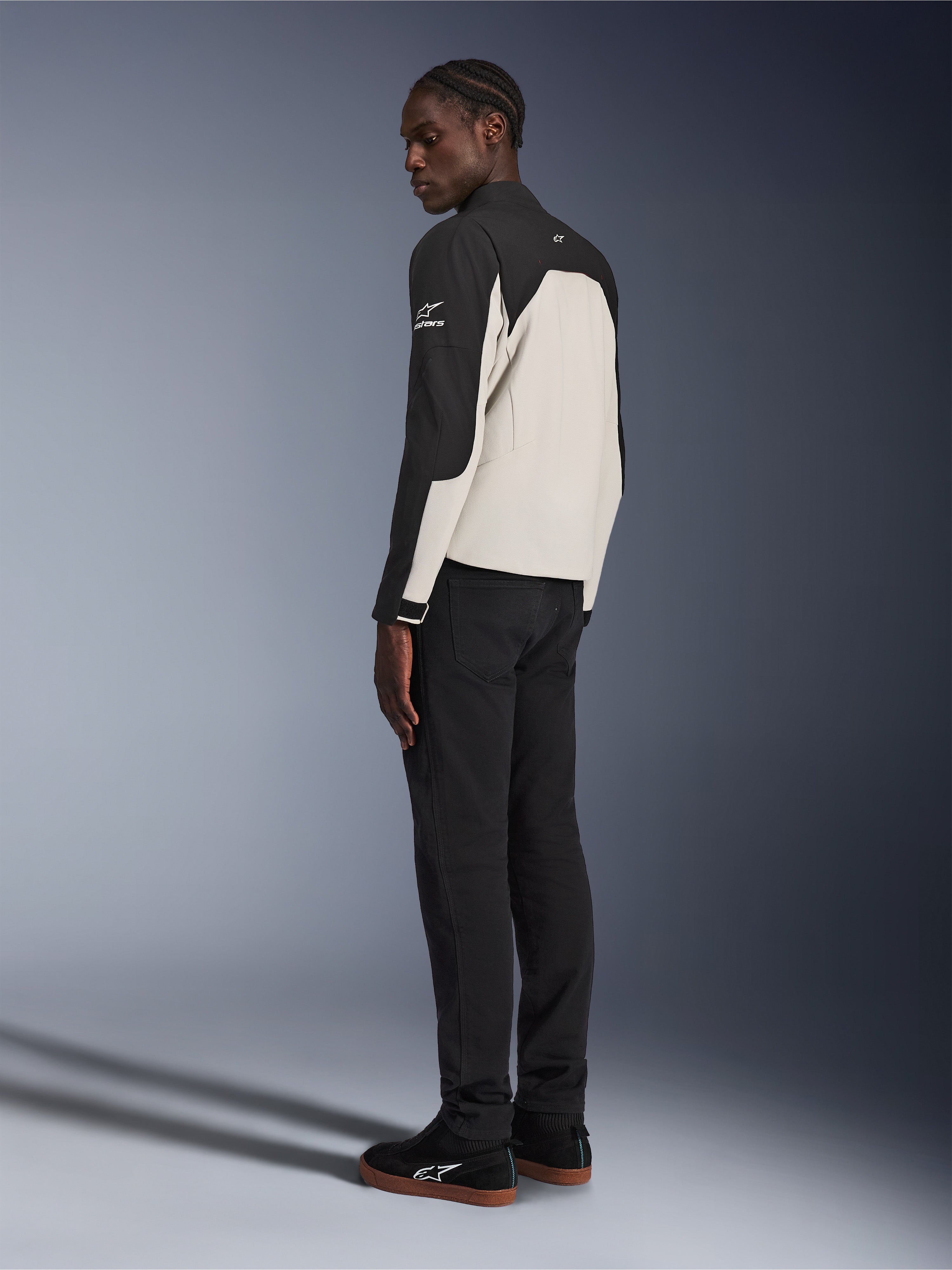 Alpinestars Provoke Jas, stedelijk motorjack, Zwart en Metal Gray, driekwart achteraanzicht op een model, tweekleurige textielconstructie met Alpinestars-logo op de schouder, gedragen met zwarte broek en zwarte sneakers, eigentijdse straatklare bescherming