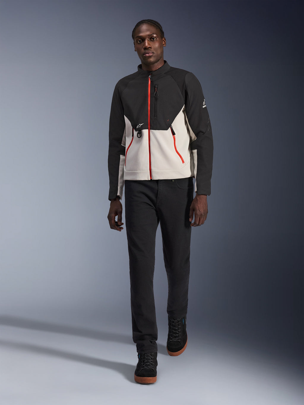 Mannelijk model draagt het Alpinestars Provoke Jacket, een stedelijk motorjack in zwart en metaalgrijs met rode ritsaccenten, gecombineerd met een zwarte broek en zwarte hoge schoenen, moderne straatklare beschermende kleding voor stadsgebruik