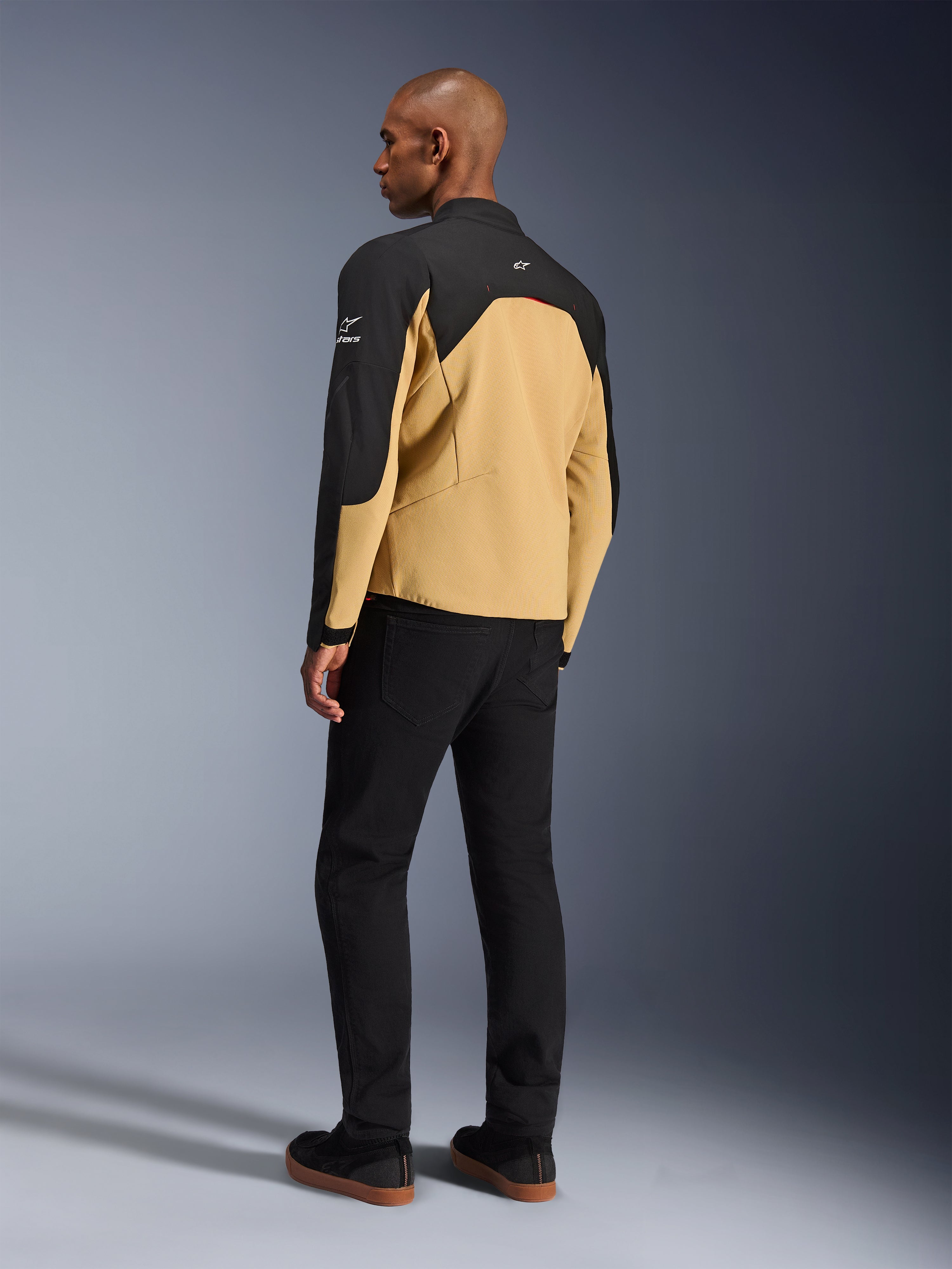 Alpinestars Provoke Jacket, urban motorjas in Black Camel, driekwart achteraanzicht, camelkleurige body met zwarte schouder- en mouwpanelen, witte Alpinestars-logo's, gedragen met zwarte broek, eigentijdse technische motoruitrusting