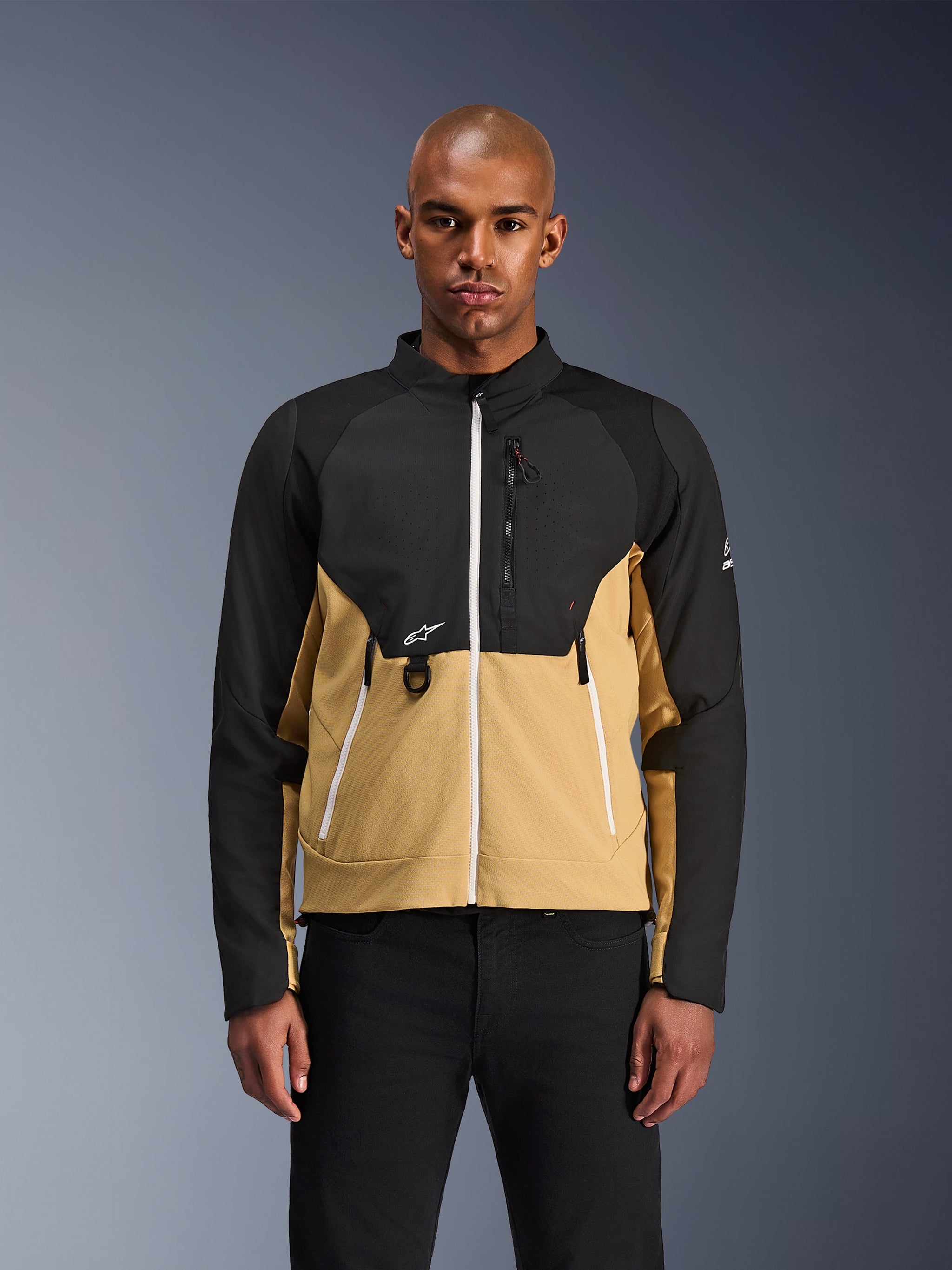 Alpinestars Provoke Jas, urban motorjas, zwart en camel, vooraanzicht op een mannelijk model, tweekleurig ontwerp met witte ritsen, borstzak met rits en D-ring en klein wit Alpinestars-logo, eigentijdse motorkleding voor de stad.