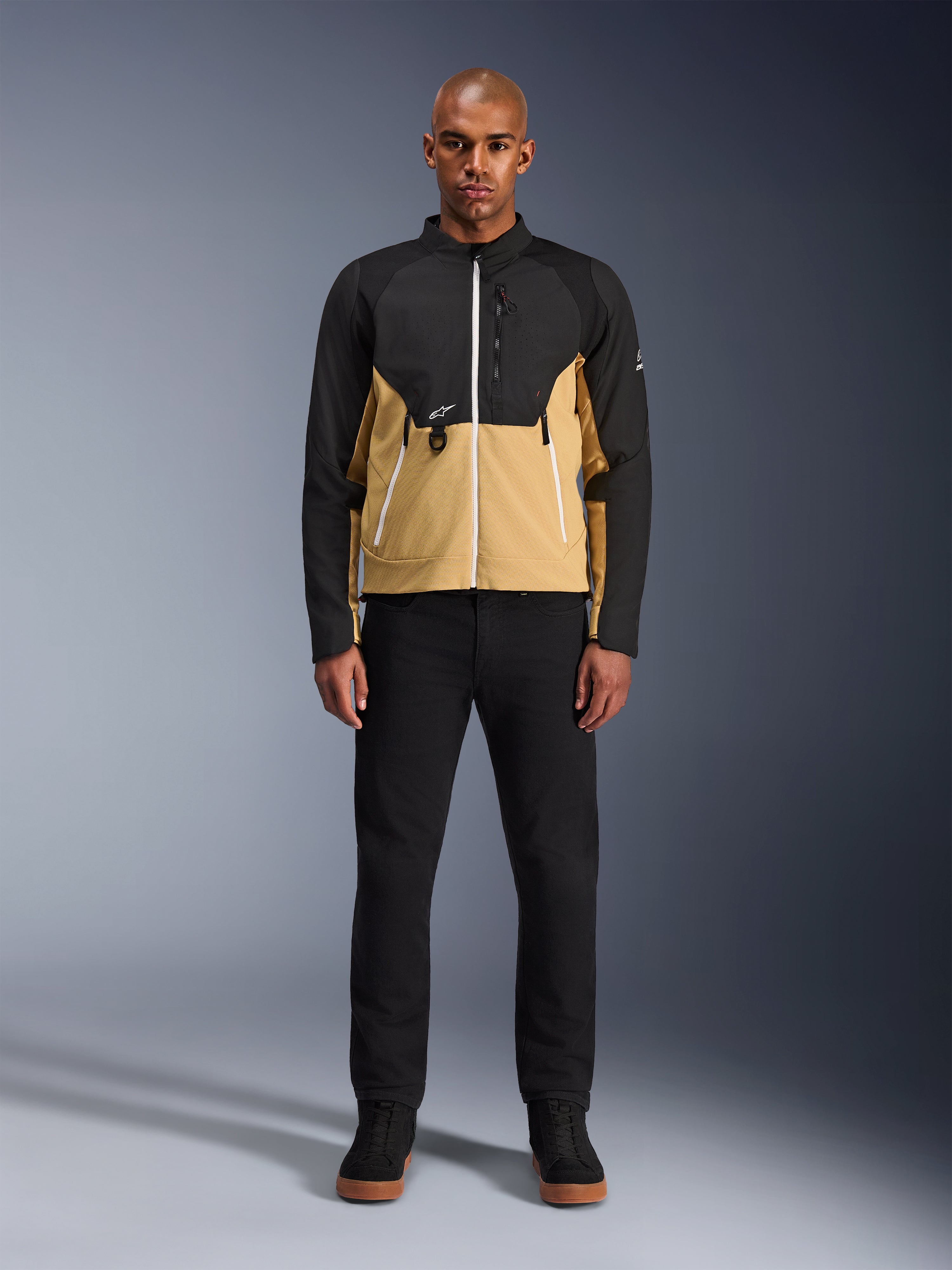 Alpinestars Provoke Jacket in Black Camel, urban motorjas met een tweekleurig zwart en camel ontwerp, gedragen door een model met zwarte broek en zwarte schoenen, eigentijds silhouet met witte ritsen en technische softshell-constructie.