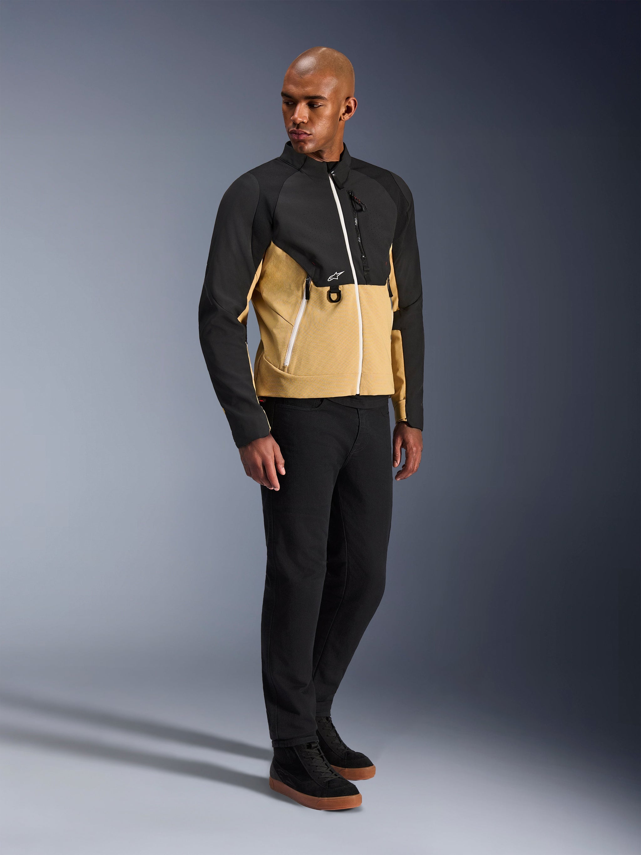 Alpinestars Provoke Jacket, urban motorjas, Black Camel, gedragen door een model met een zwarte broek en sneakers, tweekleurig textielontwerp met een zwarte bovenkant en een camelkleurig onderste deel van de romp, witte ritsdetails, eigentijdse urban motorkleding