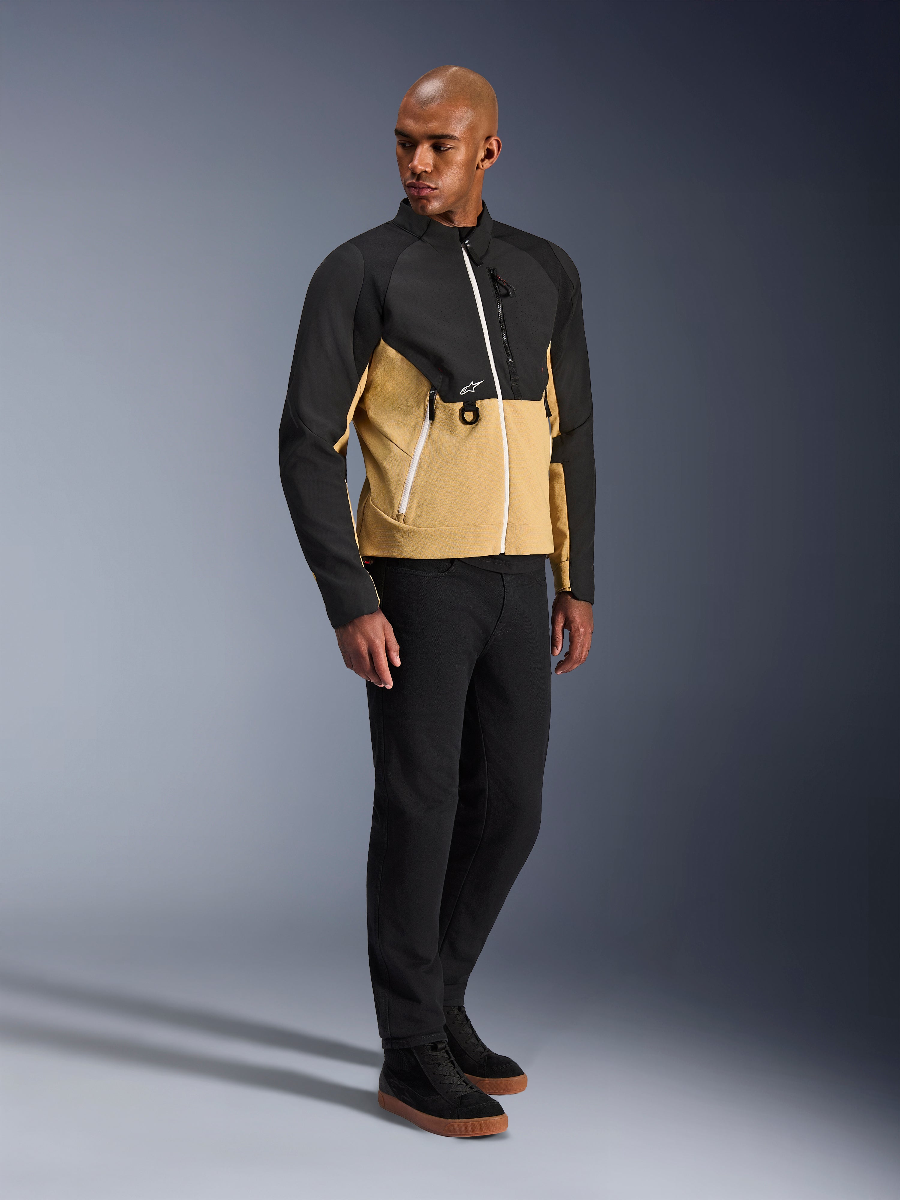 Alpinestars Provoke Jacket, urban motorjas, Black Camel, gedragen door een model met een zwarte broek en sneakers, tweekleurig textielontwerp met een zwarte bovenkant en een camelkleurig onderste deel van de romp, witte ritsdetails, eigentijdse urban motorkleding