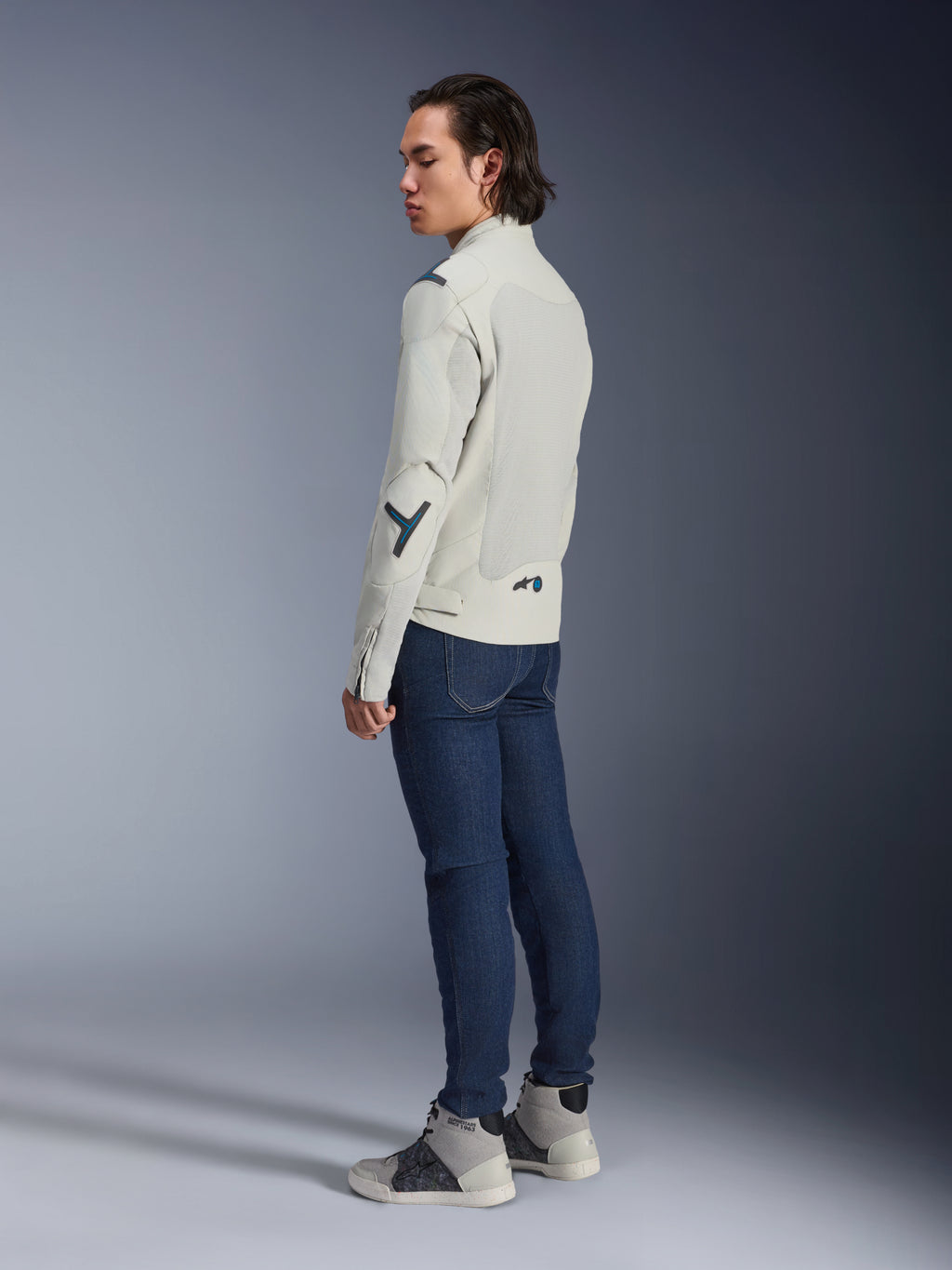 Alpinestars Circal-1 Air Jacket, stedelijke motorjas, Metal Gray met Methyl Blue accenten, gedragen door een model vanuit een driekwart achteraanzicht met donkerblauwe jeans en grijze hoge sneakers, voorzien van mesh panelen en een minimalistisch ontwerp