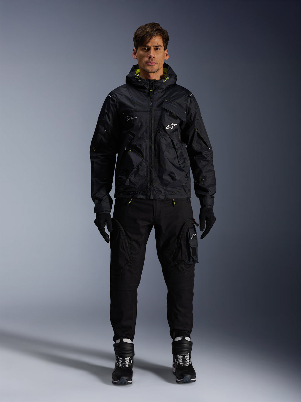 Male model wearing Alpinestars Mogress Air Jacket, black hooded textile jacket with lime punch accents, paired with black utility pants, black gloves, and black and white riding boots, urban motorsport-inspired streetwear design