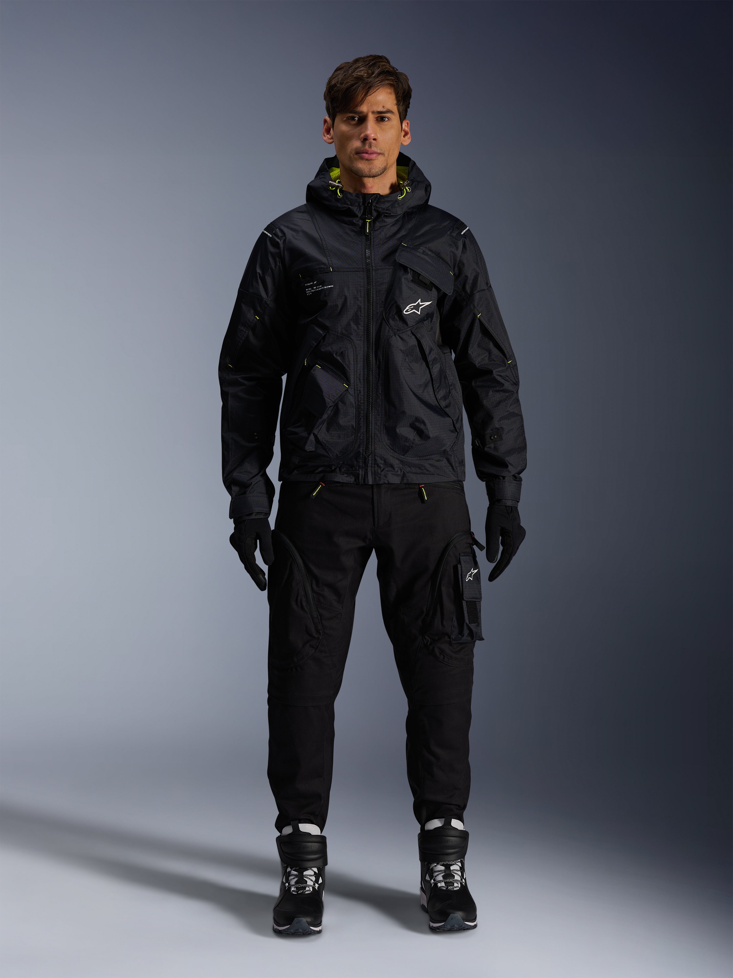 Male model wearing Alpinestars Mogress Air Jacket, black hooded textile jacket with lime punch accents, paired with black utility pants, black gloves, and black and white riding boots, urban motorsport-inspired streetwear design