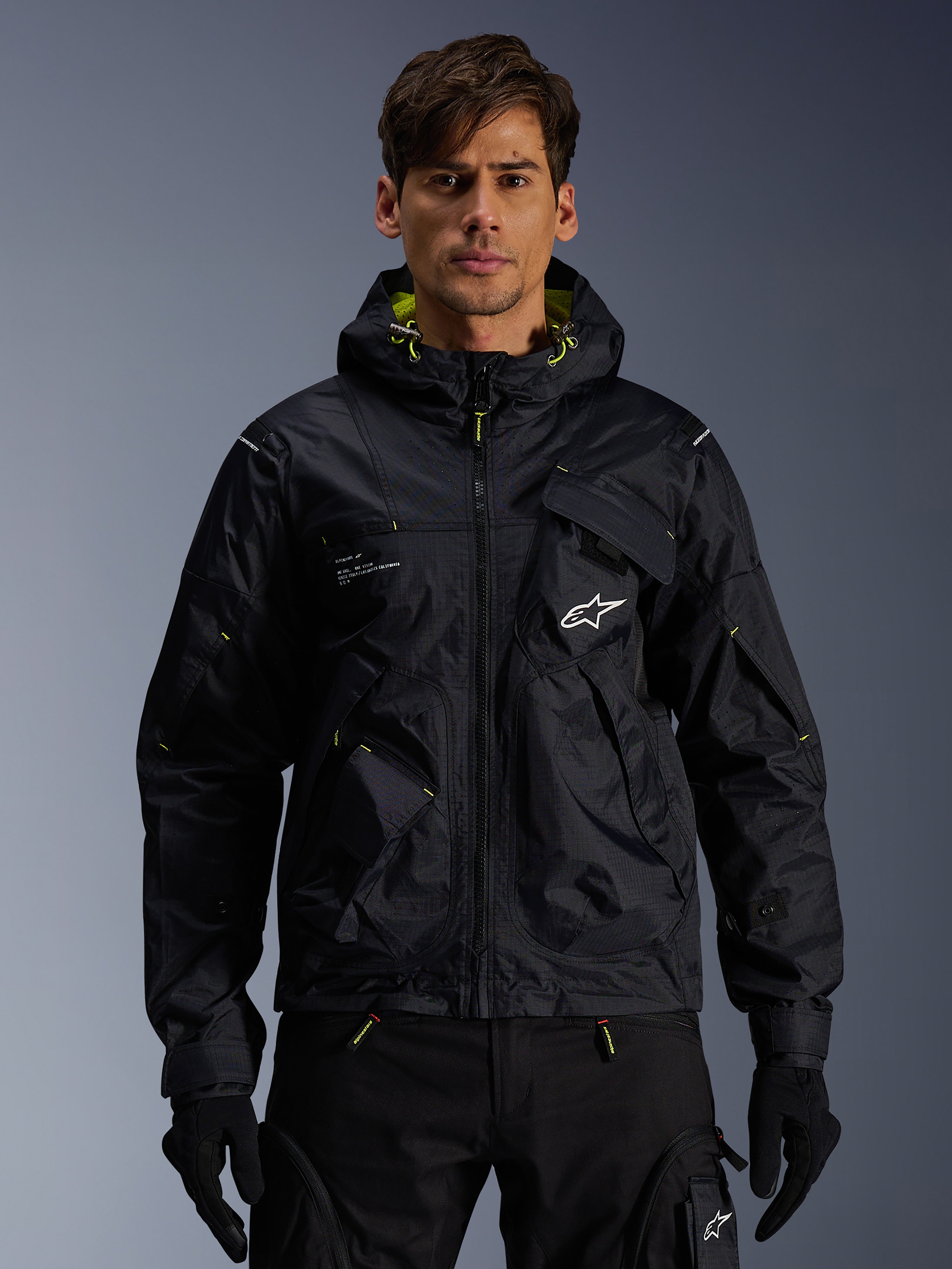 Alpinestars Mogress Air Jacket, urban motorjas, zwart met Lime Punch-accenten, gedragen door een model met zwarte motorbroek en handschoenen, op utility geïnspireerd ontwerp met een capuchon en wit sterlogo op de borst, lichtgewicht textiele uitrusting voor rijden op de weg
