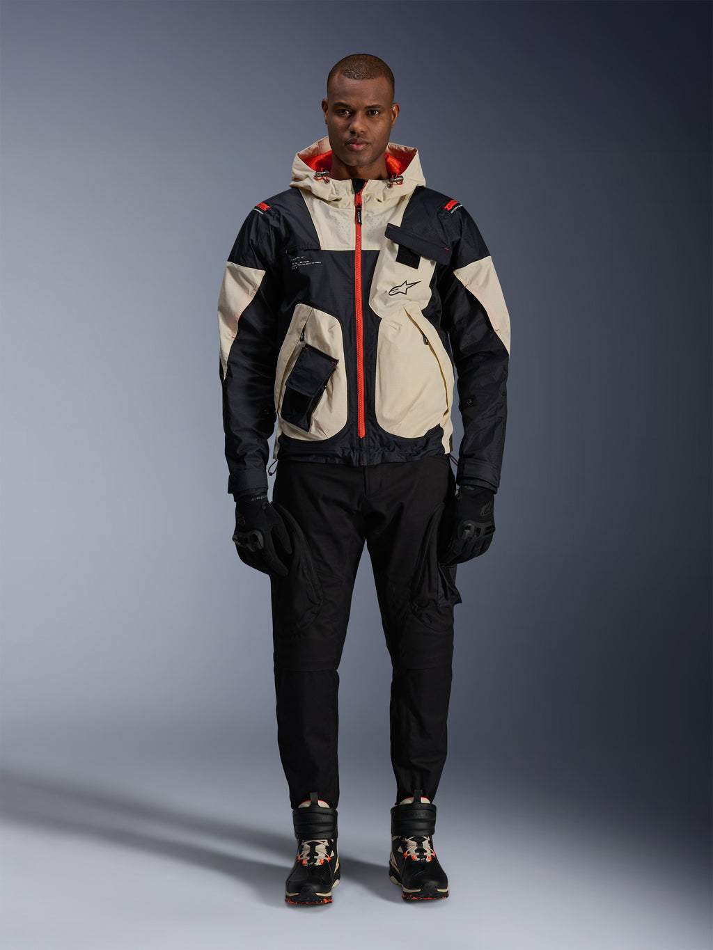 Alpinestars Mogress Air jas, urban motorjas, Black Almond en Hot Orange, gedragen door een mannelijk model met zwarte broek en handschoenen, op utility geïnspireerd ontwerp met crème panelen en feloranje ritsen, moderne streetwear beïnvloed door de motorsport.