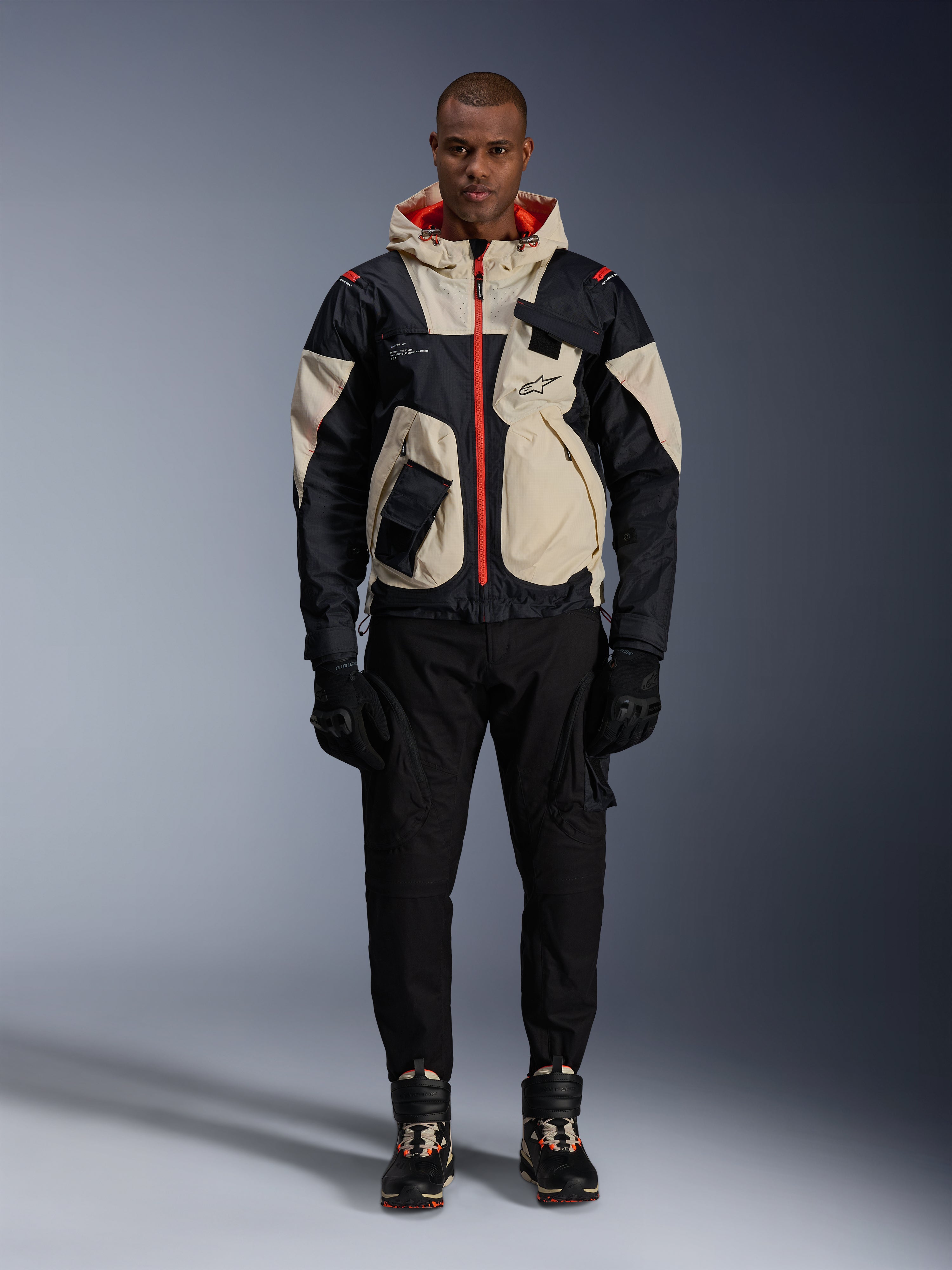 Alpinestars Mogress Air jas, urban motorjas, Black Almond en Hot Orange, gedragen door een mannelijk model met zwarte broek en handschoenen, op utility geïnspireerd ontwerp met crème panelen en feloranje ritsen, moderne streetwear beïnvloed door de motorsport.