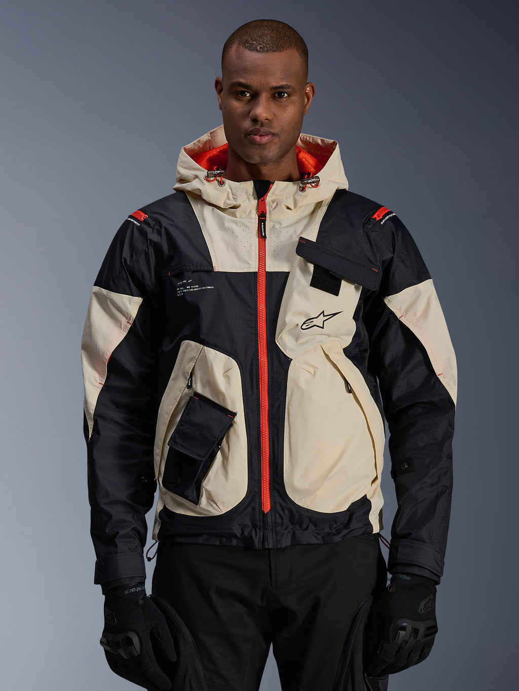 Alpinestars Mogress Air-jas, urban motorjas, Almond en zwart met Hot Orange ritsen en capuchonvoering, model met capuchon en praktische zakken, logo op de borst, gedragen met zwarte handschoenen