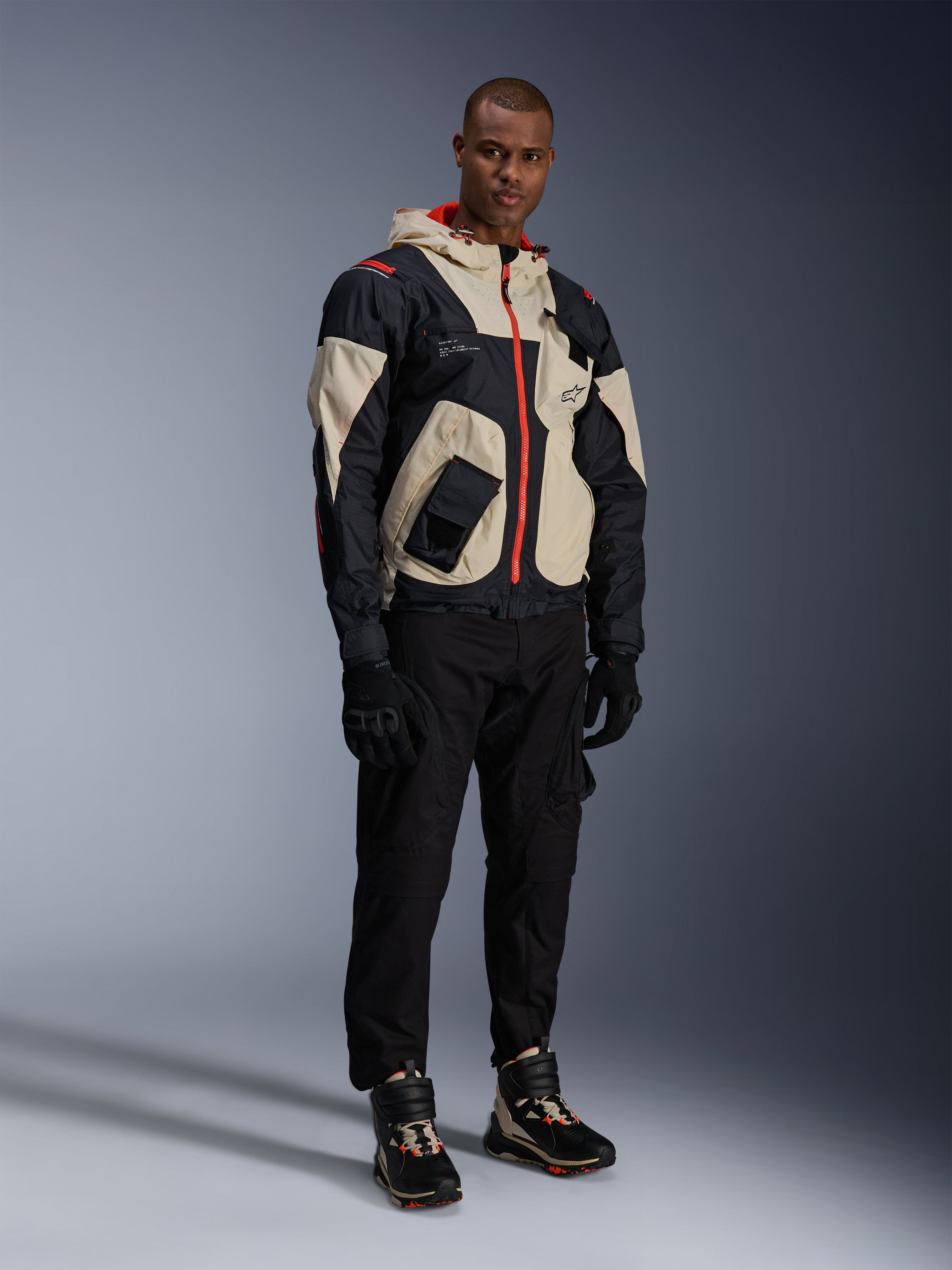 Model draagt de Alpinestars Mogress Air Jas in zwart, amandel en fel oranje, stedelijke motorjas met een utility-geïnspireerd ontwerp, gedragen met een zwarte motorbroek, zwarte handschoenen en bijpassende technische laarzen, eigentijdse street-ready motoruitrusting