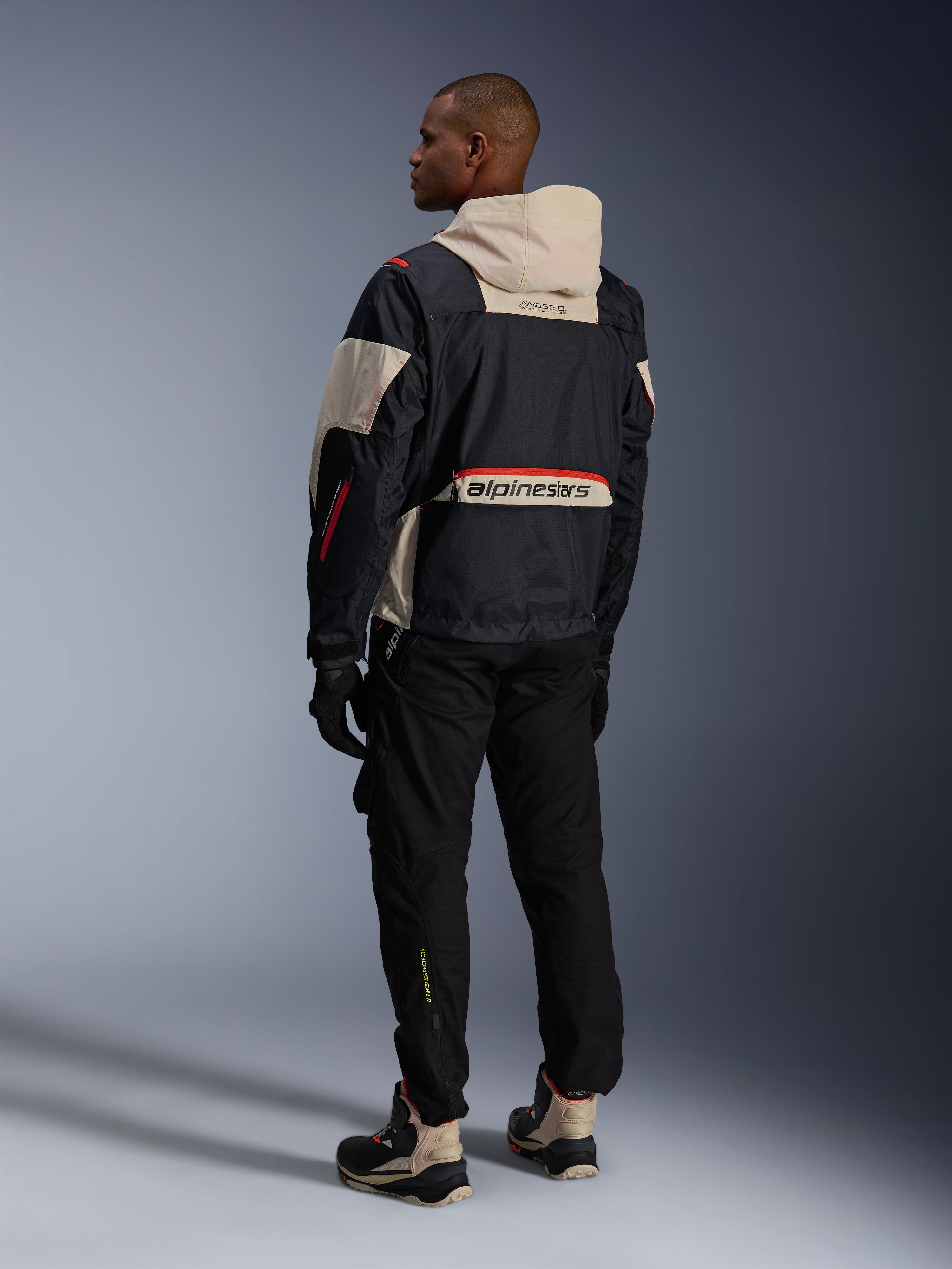 Achteraanzicht van een man die een Alpinestars Acteon broek in het zwart draagt, gecombineerd met een Mogress Air-jas in zwart en crème, zwarte handschoenen en motorlaarzen, eigentijdse urban motoroutfit