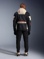Achteraanzicht van een motorrijder met de Alpinestars Mogress Air Jas in Zwart, Almond en Hot Orange, voorzien van een crème-kleurige capuchon, groot Alpinestars-logo op de achterzak en oranje ritsaccenten, gecombineerd met een zwarte broek en crème-kleurige laarzen voor een urban motorlook.