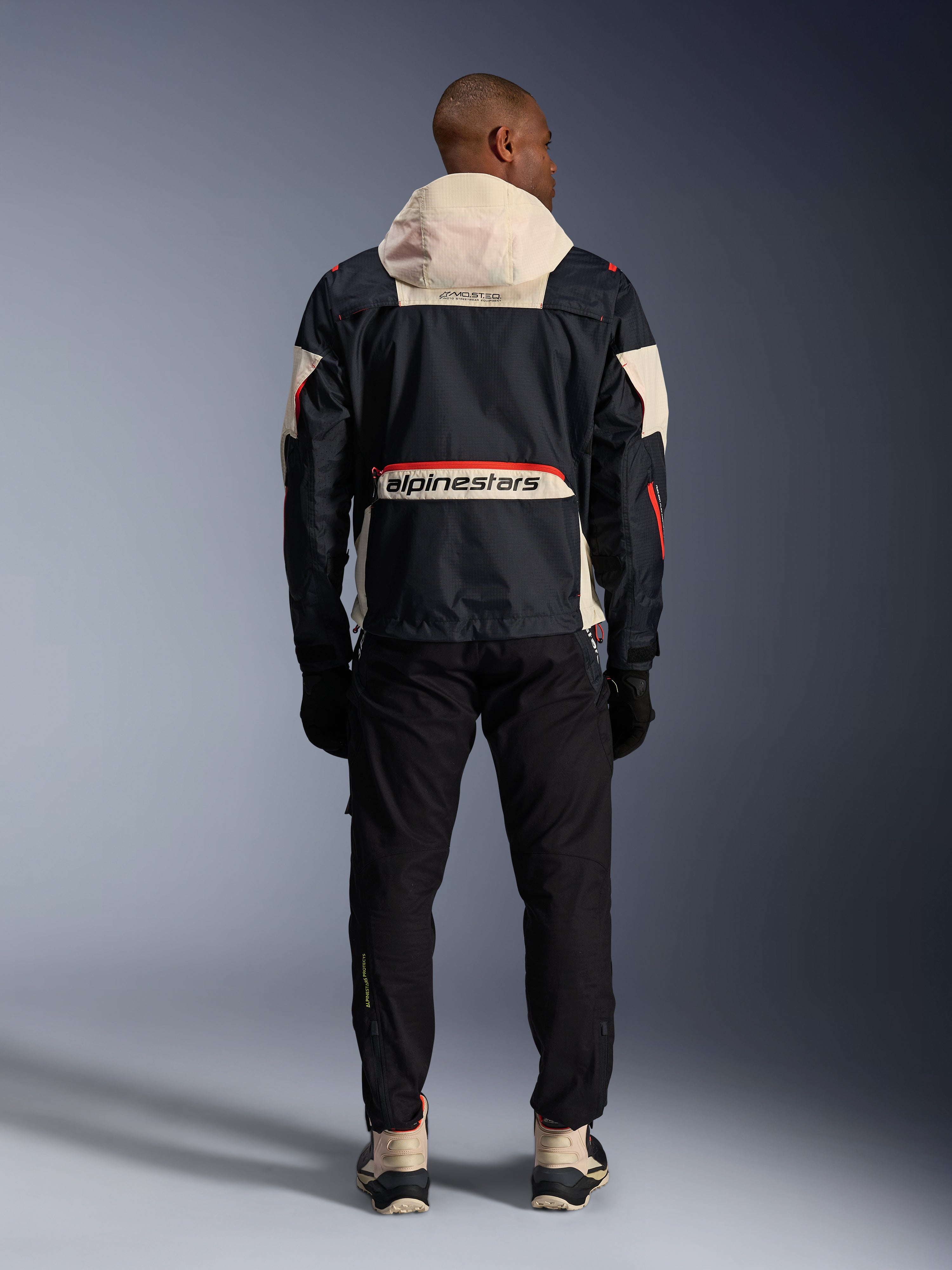 Achteraanzicht van een motorrijder met de Alpinestars Mogress Air Jas in Zwart, Almond en Hot Orange, voorzien van een crème-kleurige capuchon, groot Alpinestars-logo op de achterzak en oranje ritsaccenten, gecombineerd met een zwarte broek en crème-kleurige laarzen voor een urban motorlook.