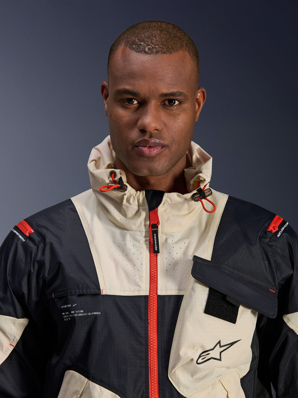 Mannelijk model draagt Alpinestars Mogress Air Jacket, urban motorjas, Black Almond en Hot Orange, close-up weergave van het color-blocked ontwerp, ripstop-textuur, capuchon met oranje stoppers en ritsdetails aan de voorzijde.