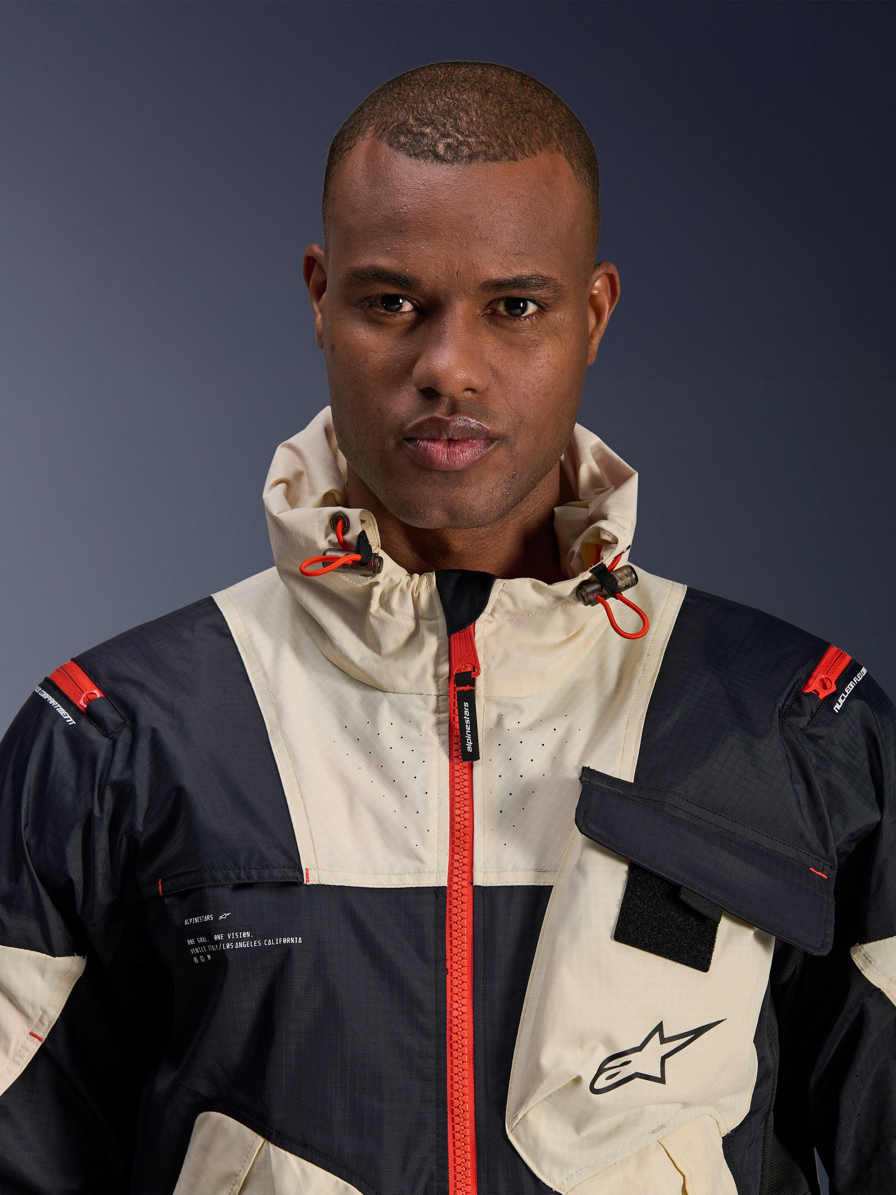Mannelijk model draagt Alpinestars Mogress Air Jacket, urban motorjas, Black Almond en Hot Orange, close-up weergave van het color-blocked ontwerp, ripstop-textuur, capuchon met oranje stoppers en ritsdetails aan de voorzijde.