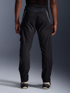 Alpinestars Acteon broek, urban motorbroek, zwart, achteraanzicht met taps toelopende pasvorm, witte merkdetails op de bovenbenen en geel logo op de kuit, verstelbare broekspijpen met ritsen, gedragen met crème sneakers
