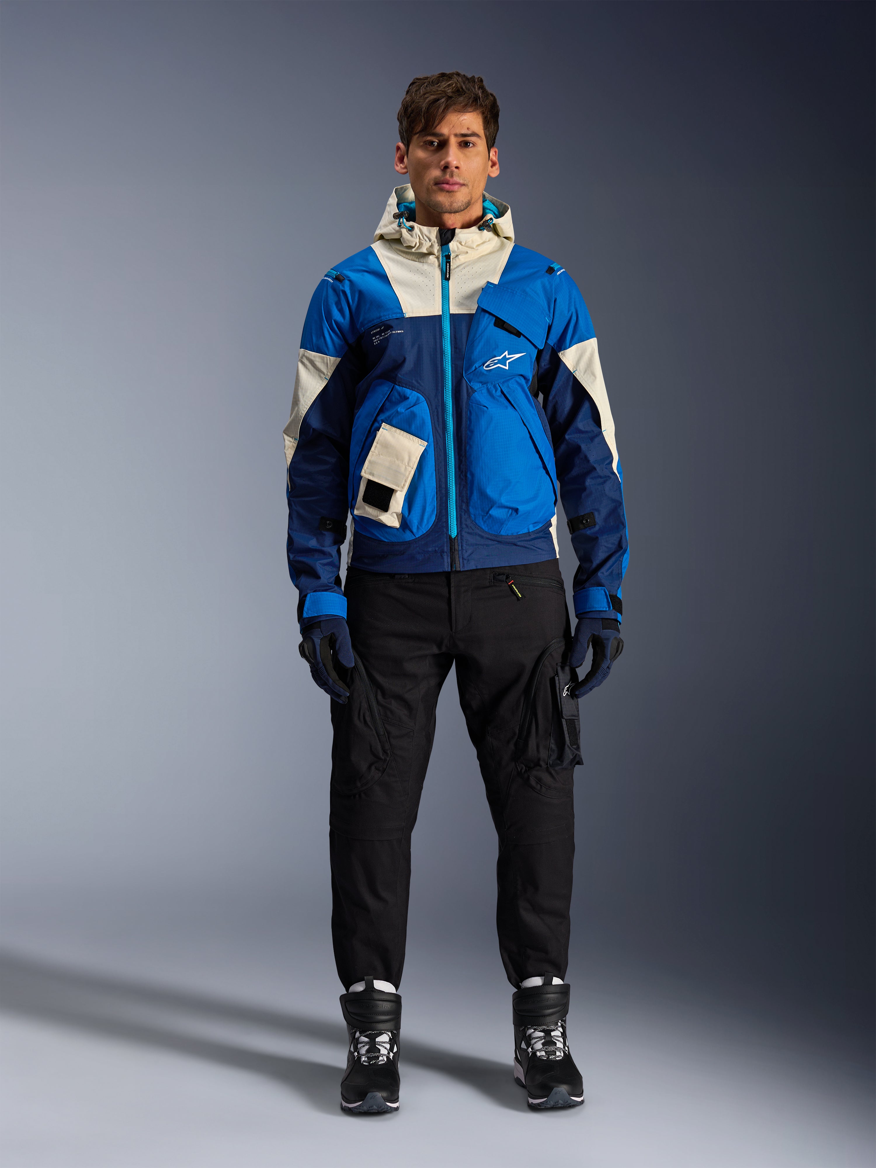Alpinestars Mogress Air Jacket, Blue Almond urban motorjas, gedragen met een zwarte broek en laarzen, voorzien van een color-blocked blauw en crème technisch ontwerp met utility-zakken en een capuchon, eigentijdse op streetwear geïnspireerde motorkleding