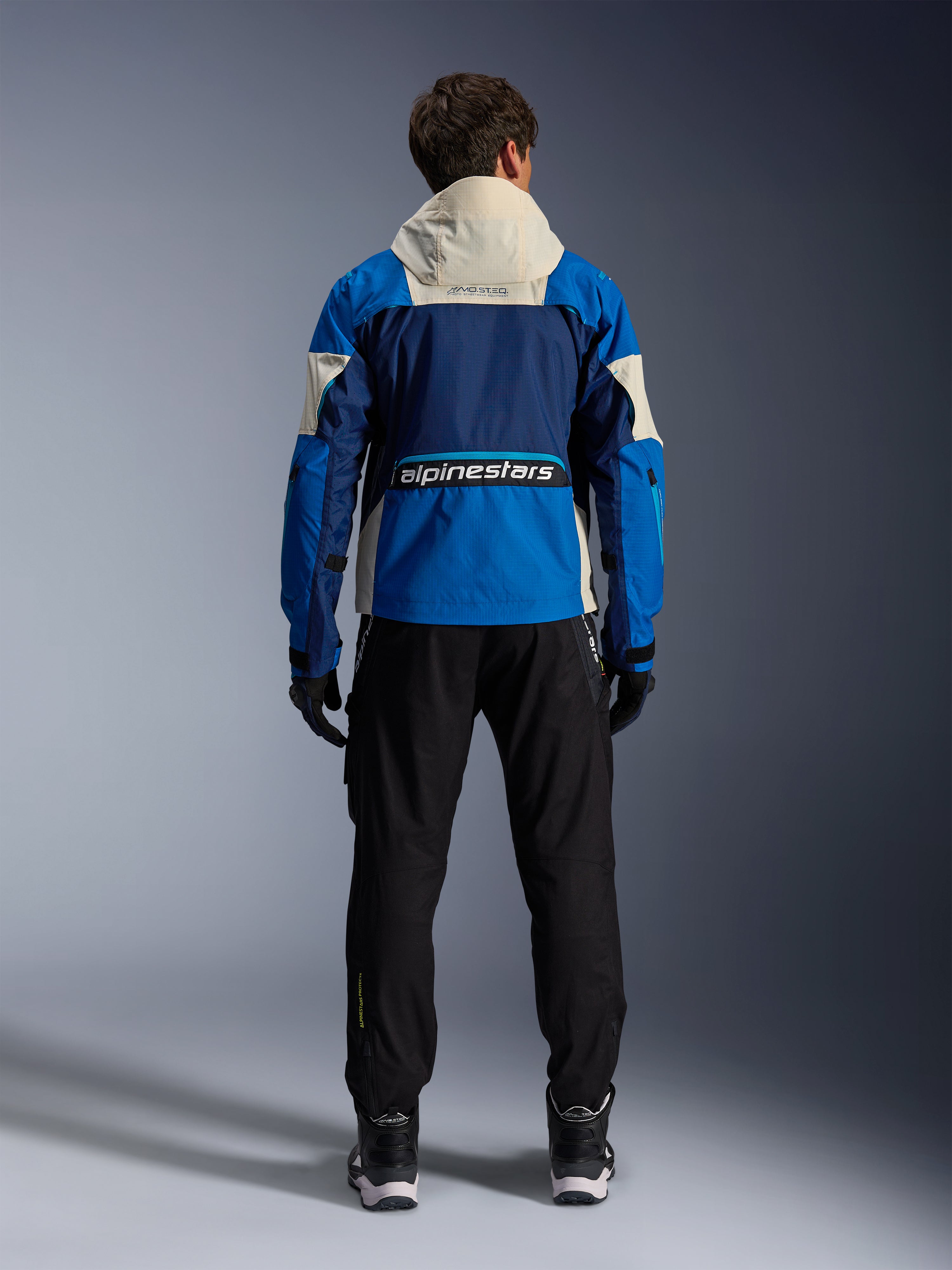 Alpinestars Mogress Air Jas in Blue Almond, achteraanzicht van een motorrijder die de motorjas draagt, voorzien van donkerblauwe en felblauwe panelen met een gebroken witte capuchon, opvallend Alpinestars-logo op de onderrug, urban contemporary kledingstijl.