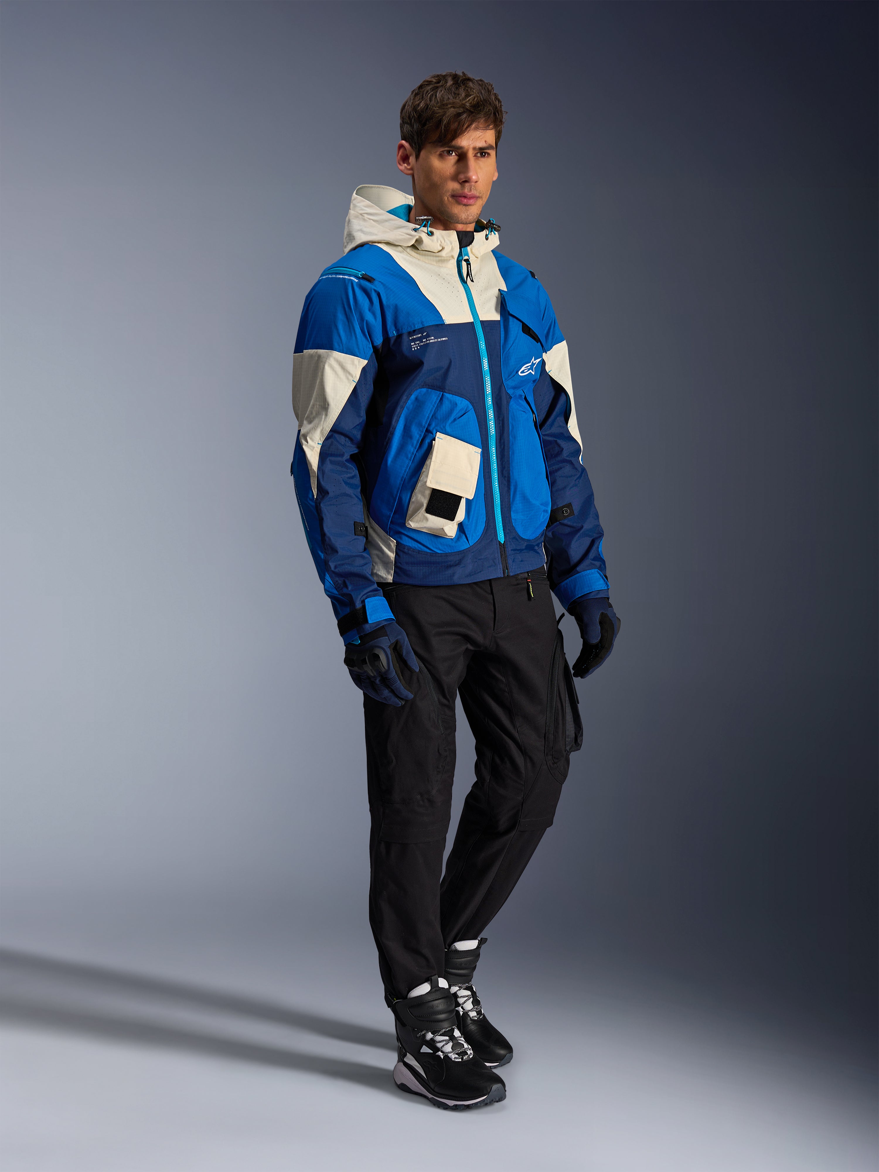 Alpinestars Mogress Air-jas, urban motorjas, Blue Almond met crème en navy panelen, voorzien van een capuchon en utility-zakken, gedragen met een zwarte motorbroek, handschoenen en laarzen, eigentijdse op streetwear geïnspireerde beschermende kleding.