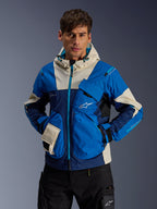 Mannelijk model draagt de Alpinestars Mogress Air-jas in Blue Almond, urban motorjas met felblauwe en marineblauwe kleurvlakken, crème accenten op de capuchon en zakken, technische ripstop-stof met utility-zakken, moderne op motorsport geïnspireerde streetwear