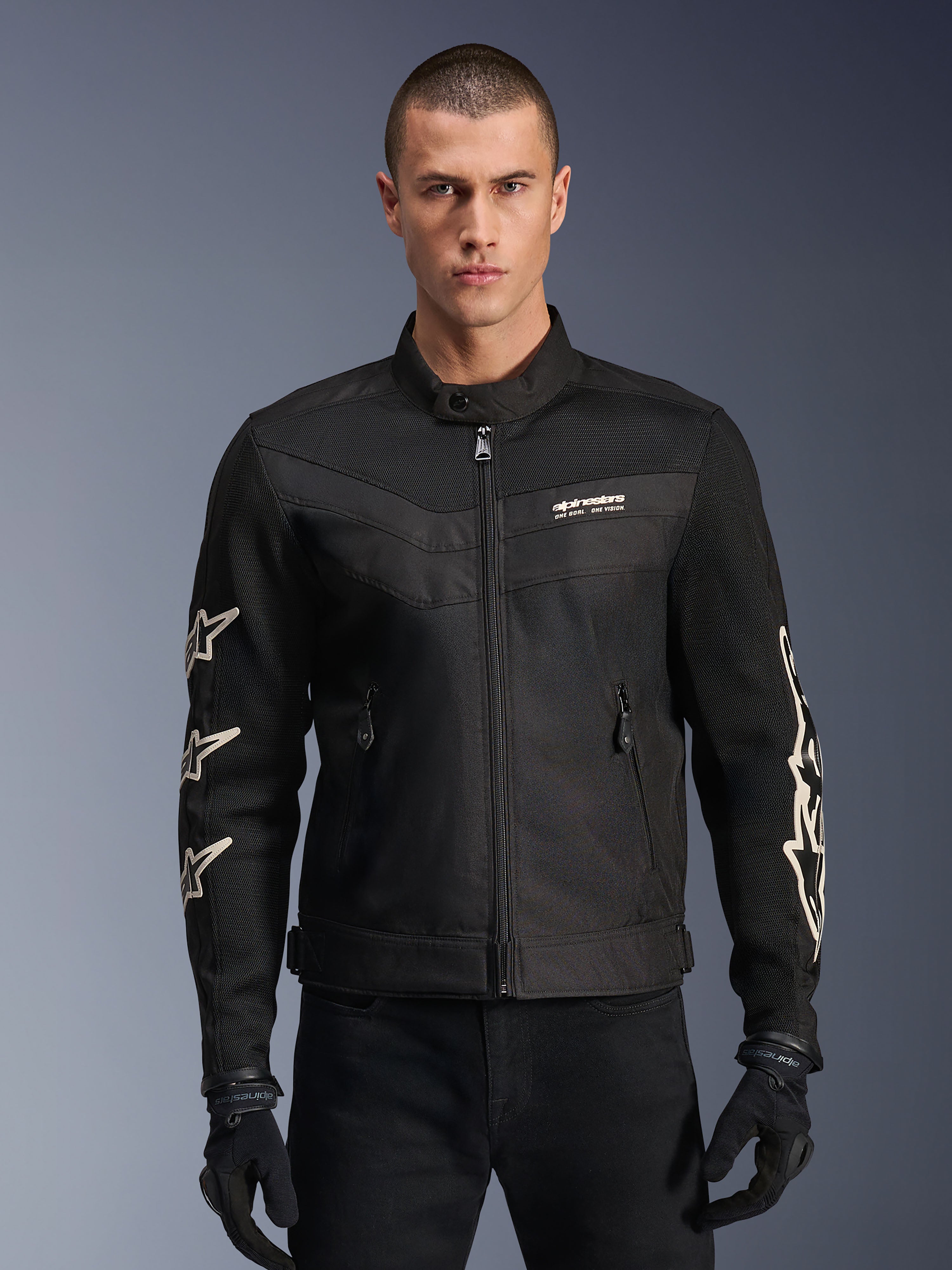 Alpinestars T-Dyno Air Jacket, zwarte textiele motorjas, voorzien van witte ster-graphics op de mouwen en logo op de borst, meshpanelen voor ventilatie, vintage-geïnspireerde biker-pasvorm, gedragen met zwarte handschoenen en broek