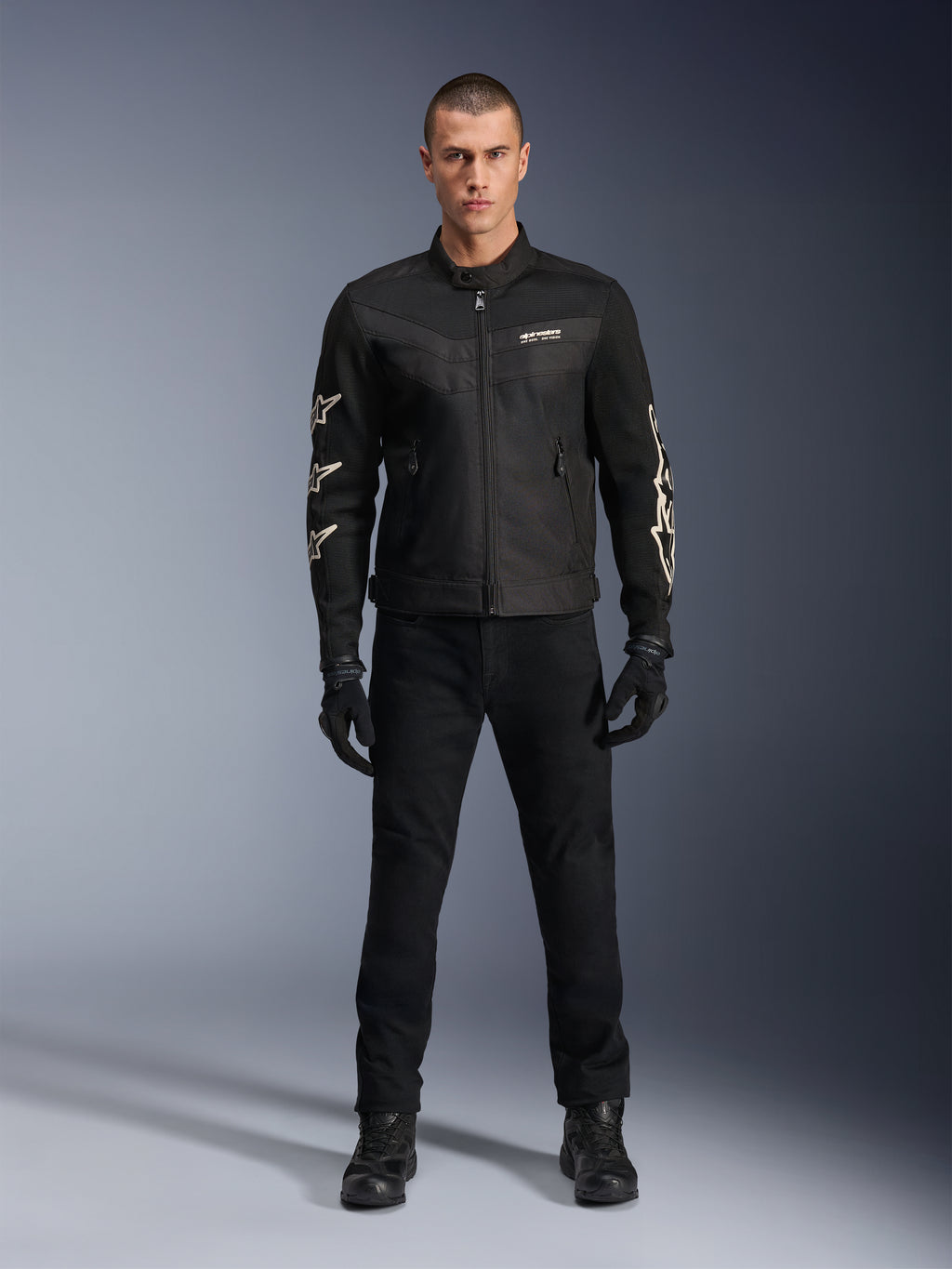 Mannelijk model in Alpinestars T-Dyno Air Jacket, zwarte lichtgewicht textiele motorjas met ademend 3D-mesh, witte sterlogo's op de mouwen, gecombineerd met zwarte broek, handschoenen en laarzen, vooraanzicht staand