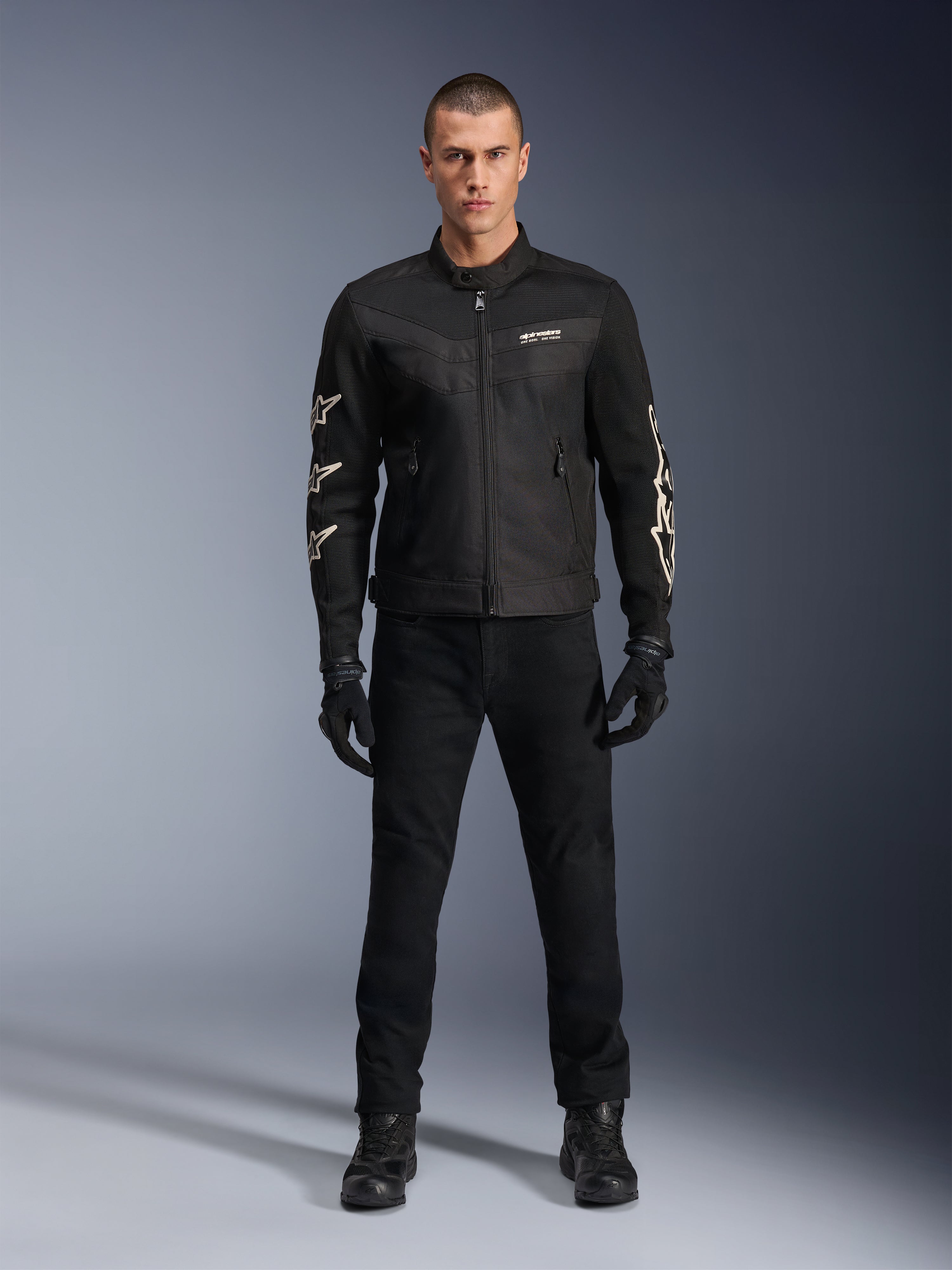 Mannelijk model in Alpinestars T-Dyno Air Jacket, zwarte lichtgewicht textiele motorjas met ademend 3D-mesh, witte sterlogo's op de mouwen, gecombineerd met zwarte broek, handschoenen en laarzen, vooraanzicht staand