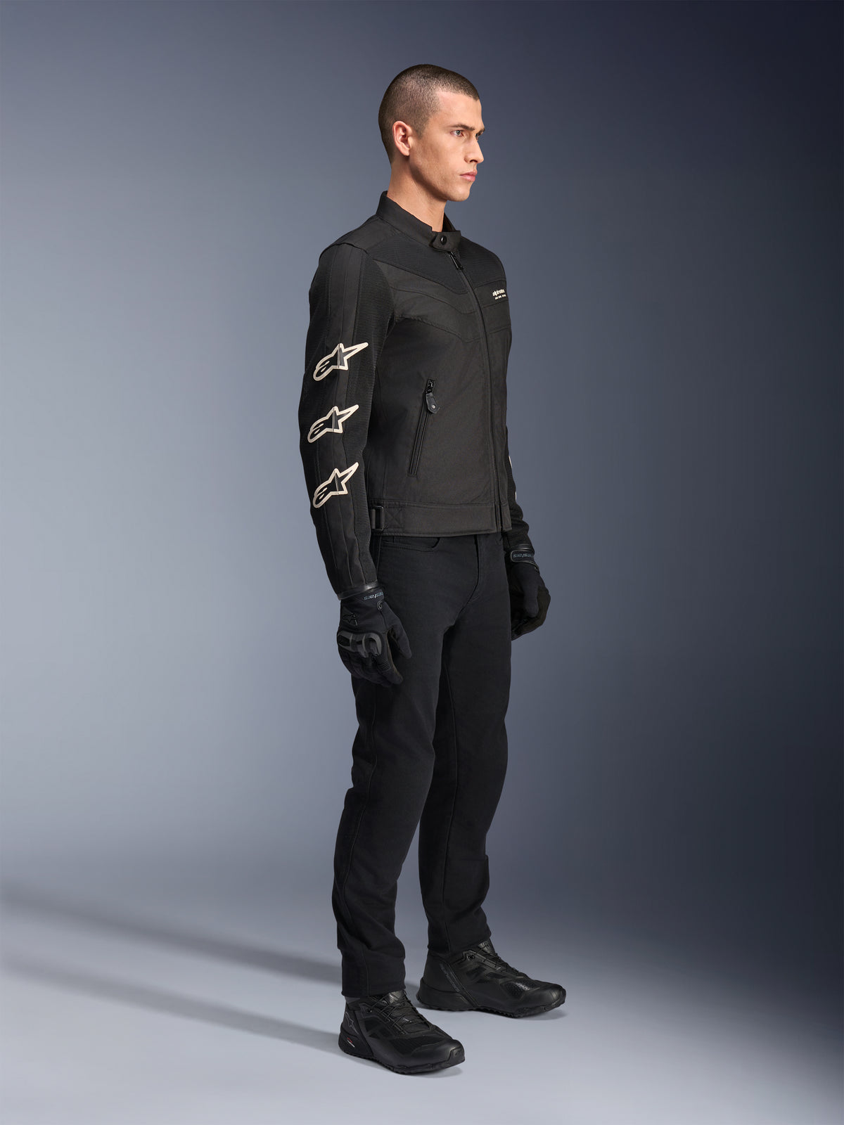 Alpinestars T-Dyno Air Jacket, zwarte textiel motorjas, voorzien van drie witte Alpinestars-logo's op de mouw, gedragen met bijpassende zwarte broek en handschoenen, urban motorkleding met ademende mesh-panelen