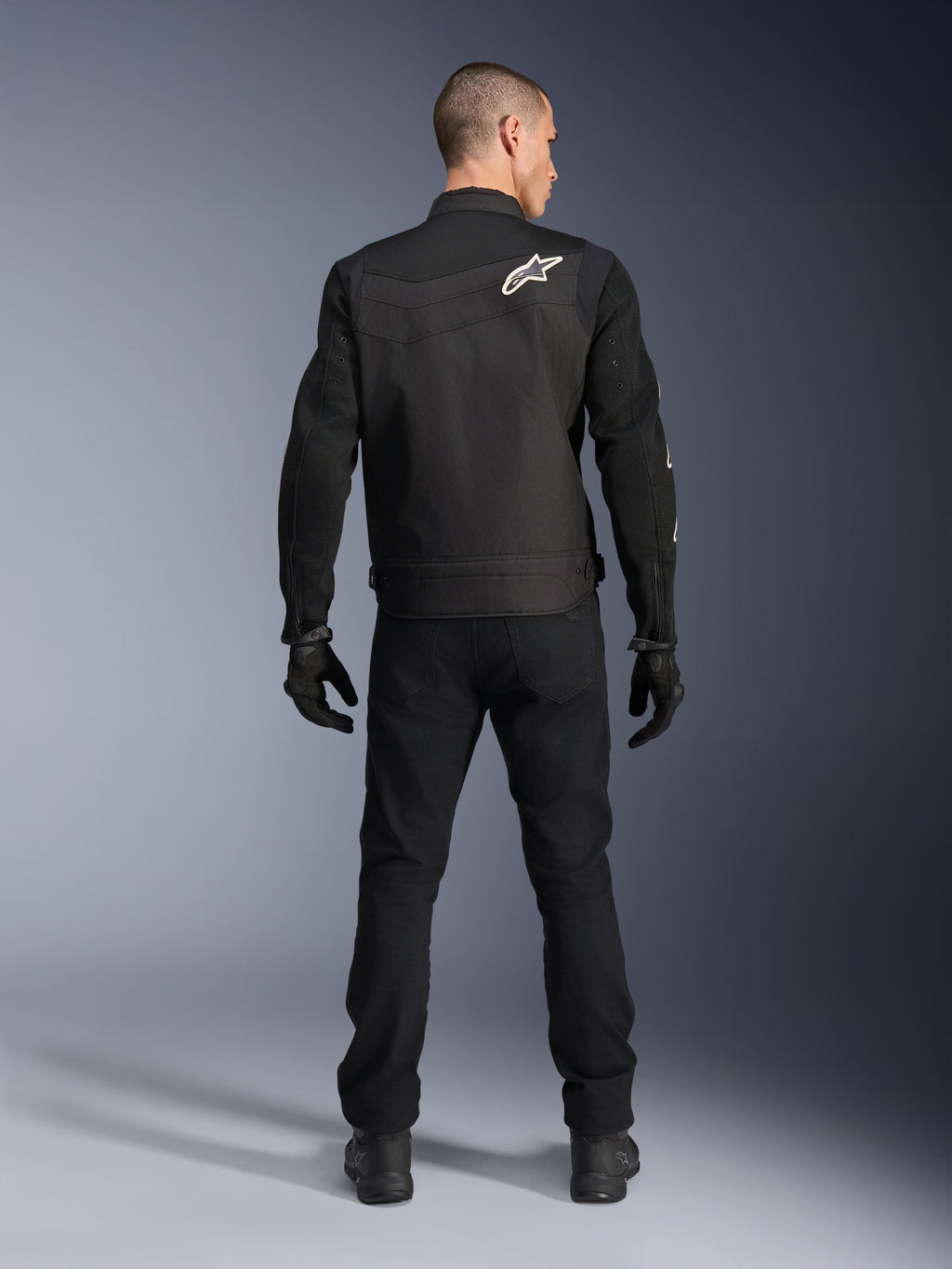 Alpinestars T-Dyno Air Jacket, zwart, achteraanzicht op model, textiel motorjas met 3D-mesh panelen en een wit sterlogo op de schouder, gedragen met zwarte broek en handschoenen, moderne urban motoroutfit