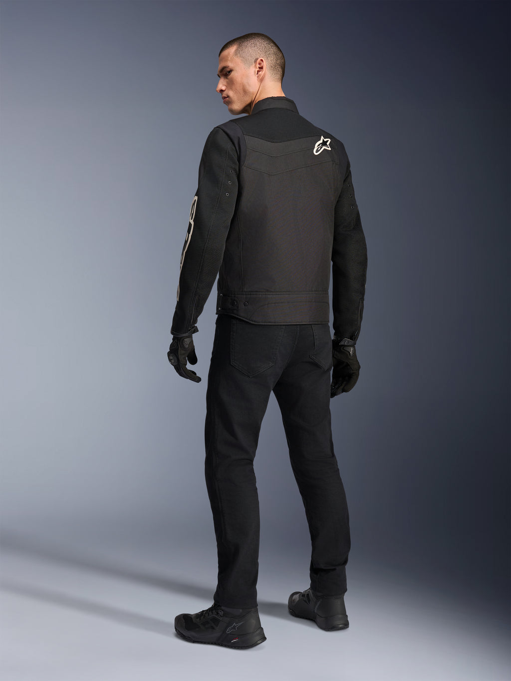 Alpinestars T-Dyno Air Jas, zwart, achteraanzicht van model met een ademende textiele motorjas, met wit Alpinestars-logo op de schouder, gecombineerd met een zwarte broek en handschoenen, urban rijstijl