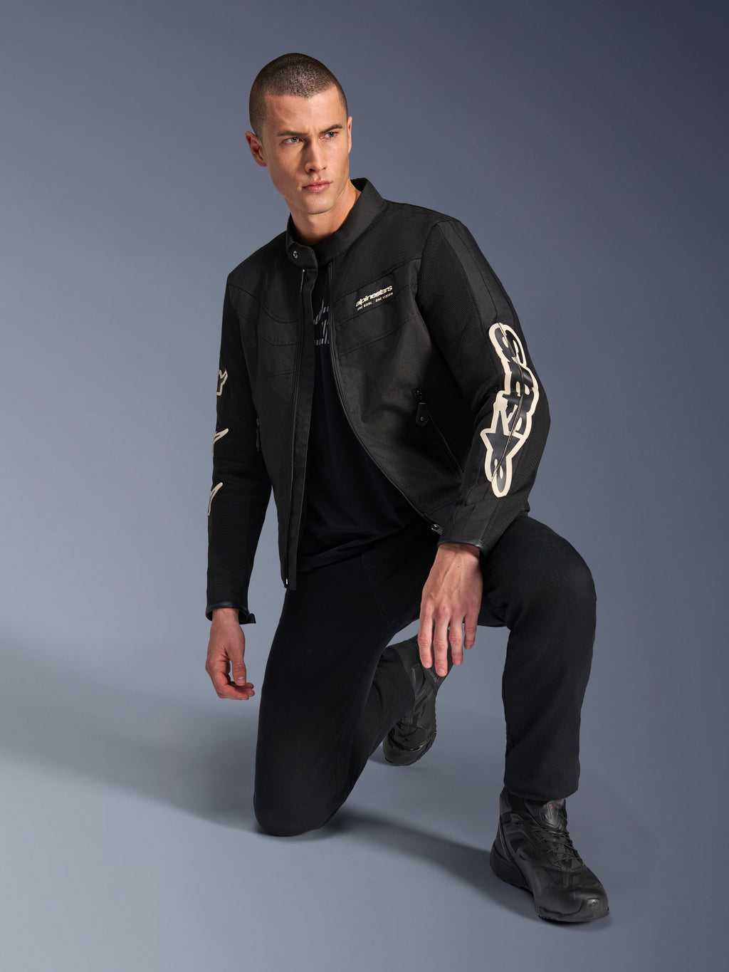 Mannelijk model draagt Alpinestars T-Dyno Air Jacket, zwarte textiele motorjas met grote witte branding op de mouwen, gecombineerd met zwarte broek en motorlaarzen, modern vintage-geïnspireerd ontwerp met 3D-meshpanelen.