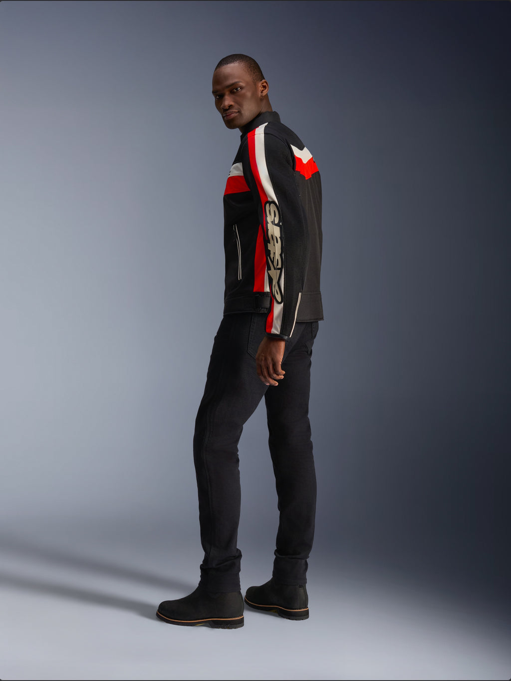 Een man die de Alpinestars T-Dyno Air Jas draagt in zwart, helderrood en ecru, vintage-geïnspireerde textiele motorjas met mesh-panelen, opvallende branding op de mouw, gecombineerd met een zwarte broek en laarzen voor een moderne biker-look
