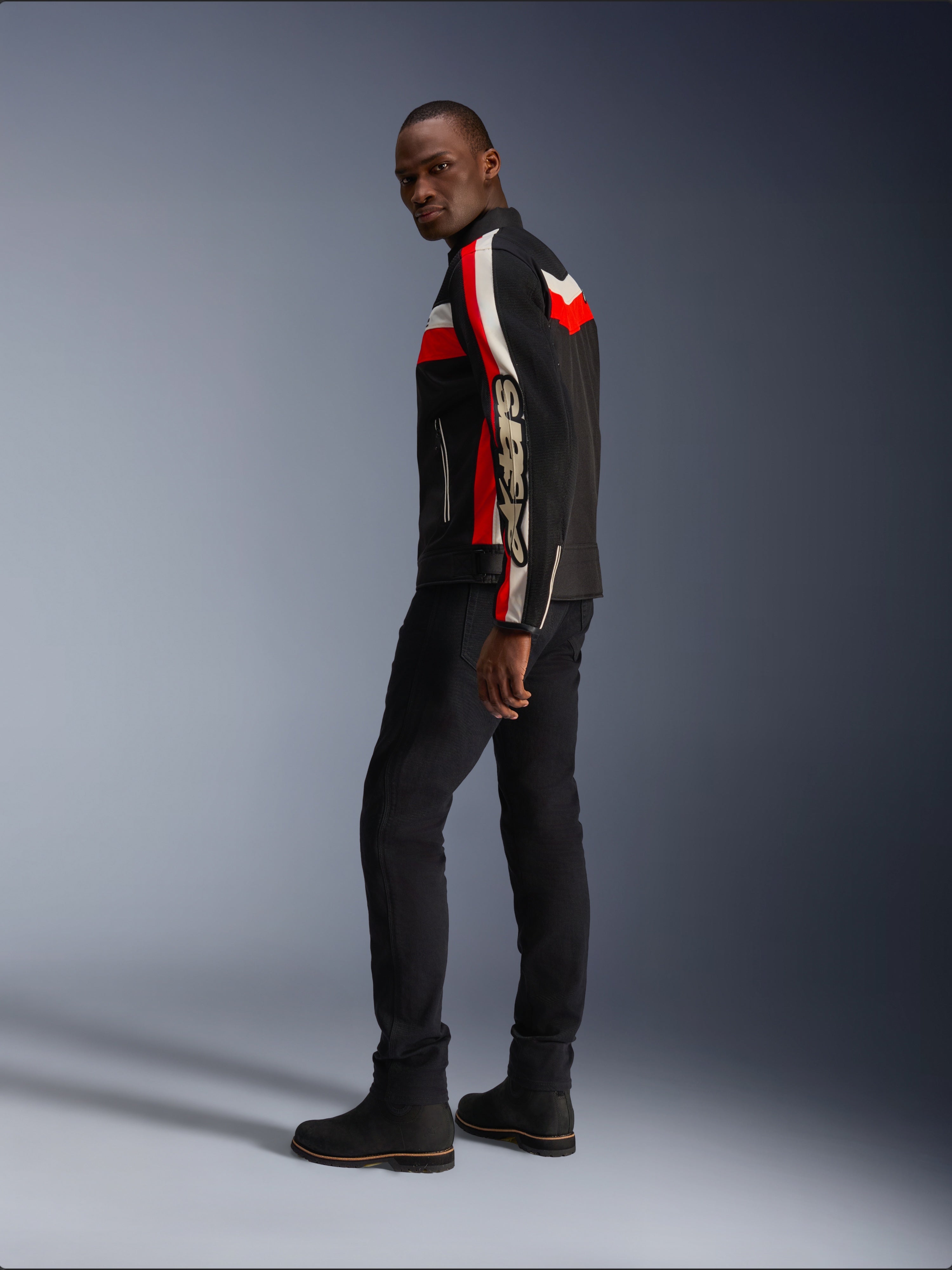 Een man die de Alpinestars T-Dyno Air Jas draagt in zwart, helderrood en ecru, vintage-geïnspireerde textiele motorjas met mesh-panelen, opvallende branding op de mouw, gecombineerd met een zwarte broek en laarzen voor een moderne biker-look
