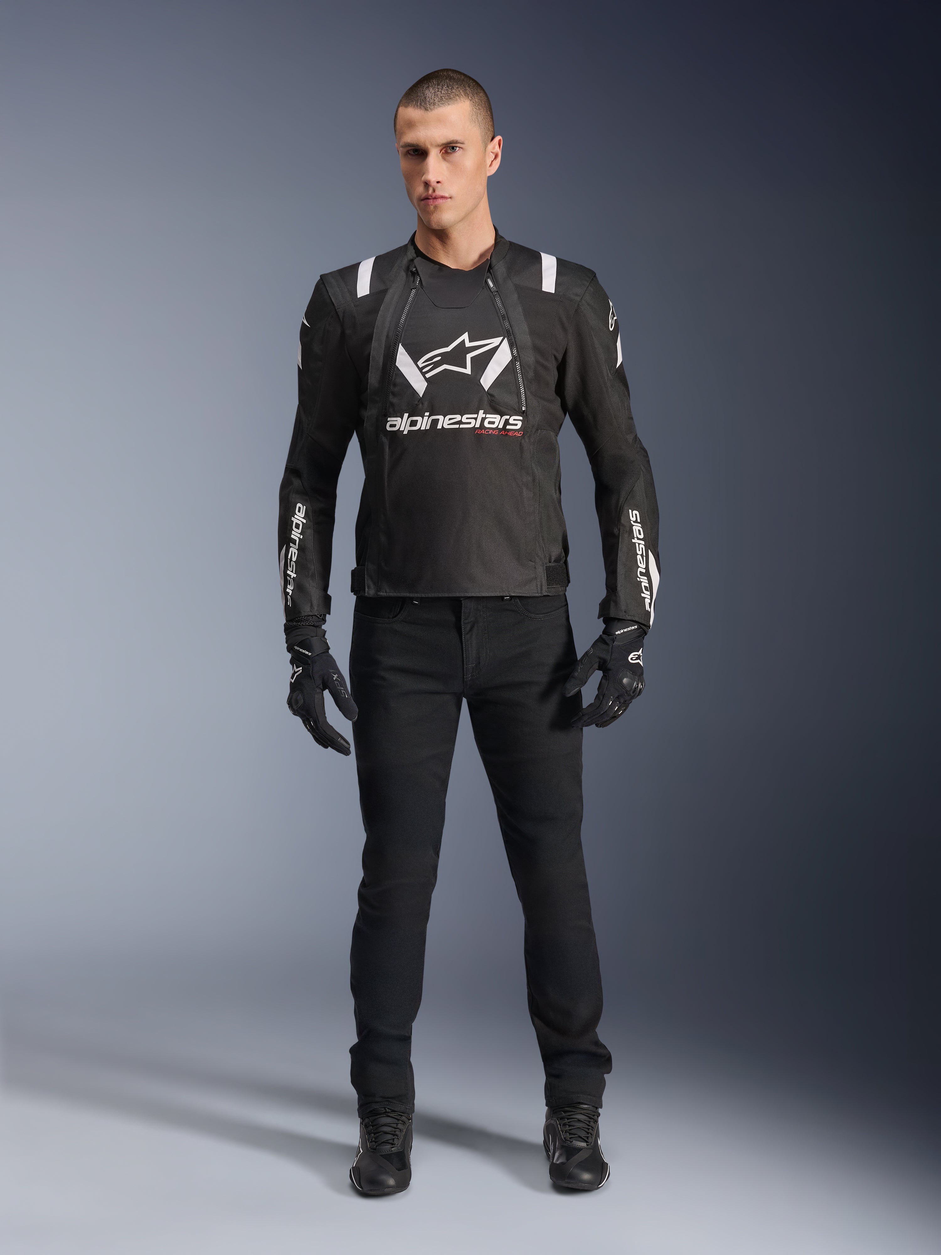Alpinestars T-Stunt Air Jas, zwart-wit motorjack, gedragen met een zwarte motorbroek, handschoenen en laarzen, voorzien van opvallende witte Alpinestars-branding op de borst en mouwen, sportief textielontwerp met mesh-panelen voor ventilatie