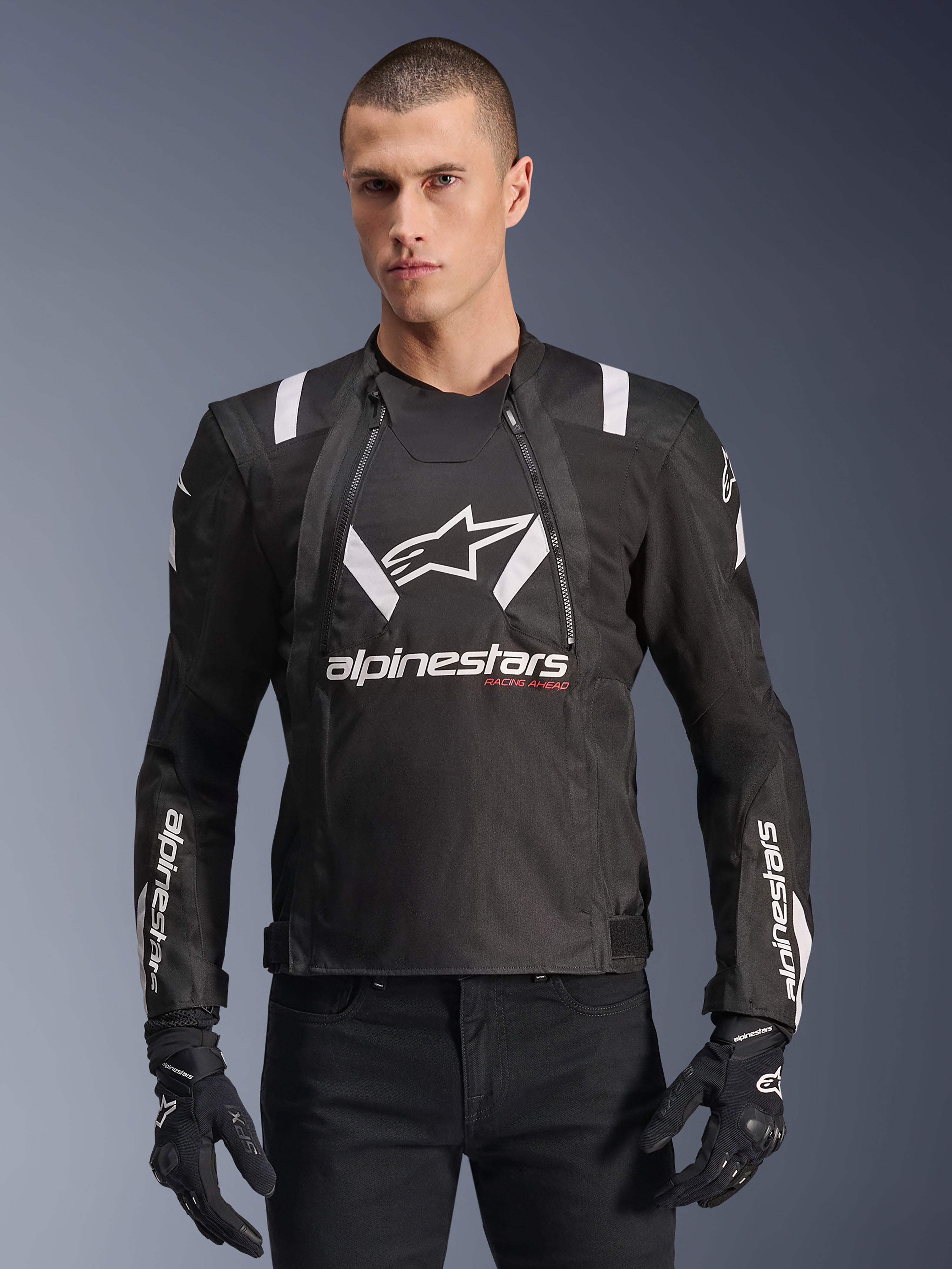 Alpinestars T-Stunt Air Jacket, zwart-witte motorjas, gedragen door een man met zwarte handschoenen en broek, met opvallende witte branding op de borst en mouwen en rode 'Racing Ahead' accenten, strak sportief design met witte schouderstrepen en ademende mesh-panelen.